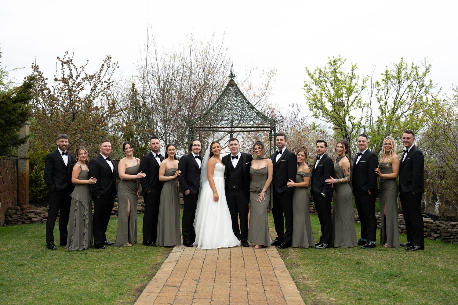 Wedding party in black tuxedos and sage green gowns pose together in garden gazebo venue.