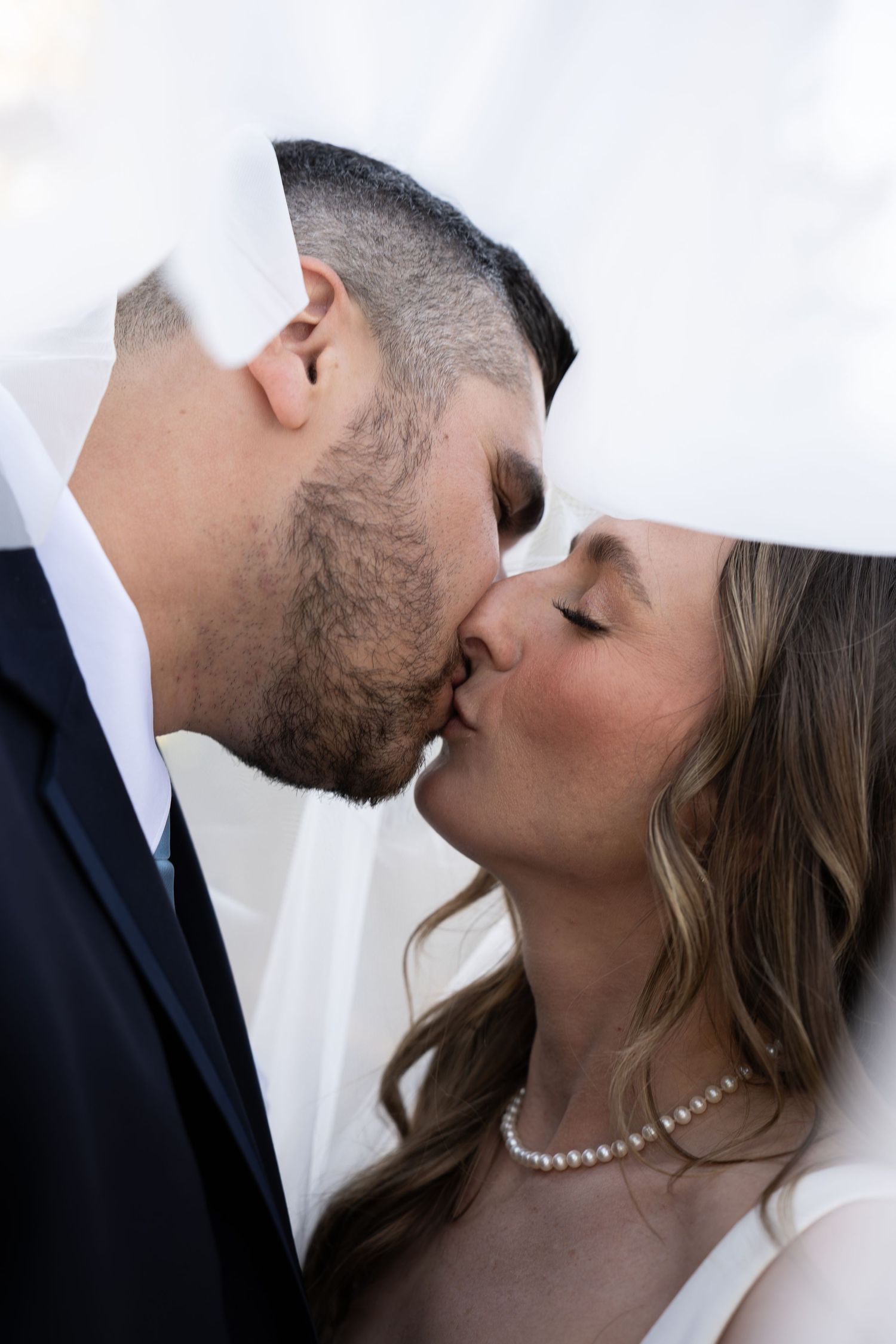 A bride and groom share a romantic kiss under a white wedding veil in a close-up portrait.