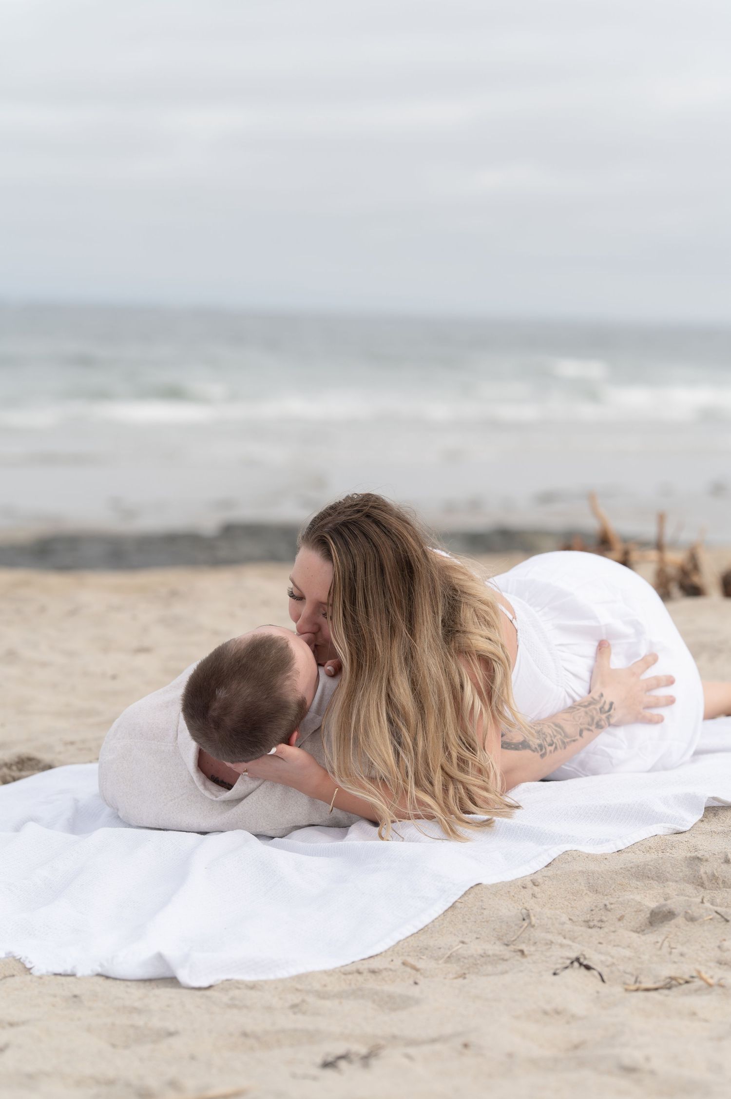 A couple shares a tender moment lying on a white blanket on a sandy beach.