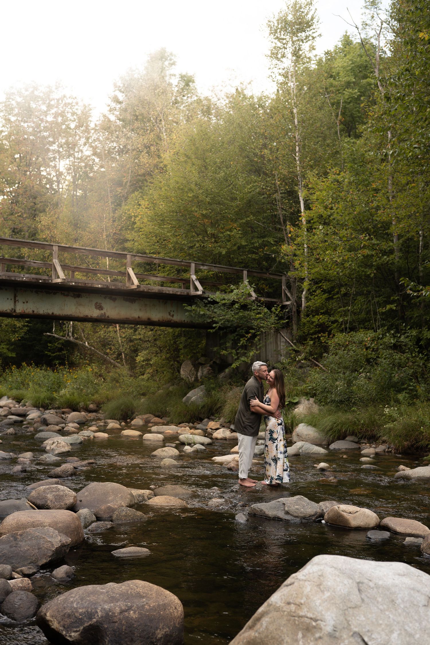 Couple embracing on rocky riverbank beneath an old bridge in misty forest.
