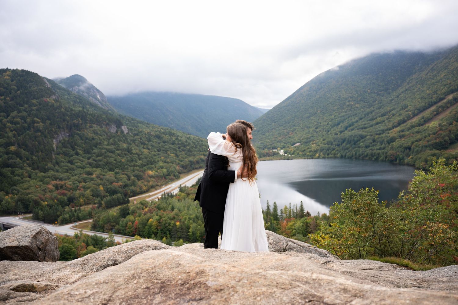 Couple embracing on rocky mountaintop overlooking scenic lake and lush green valley on their wedding day.