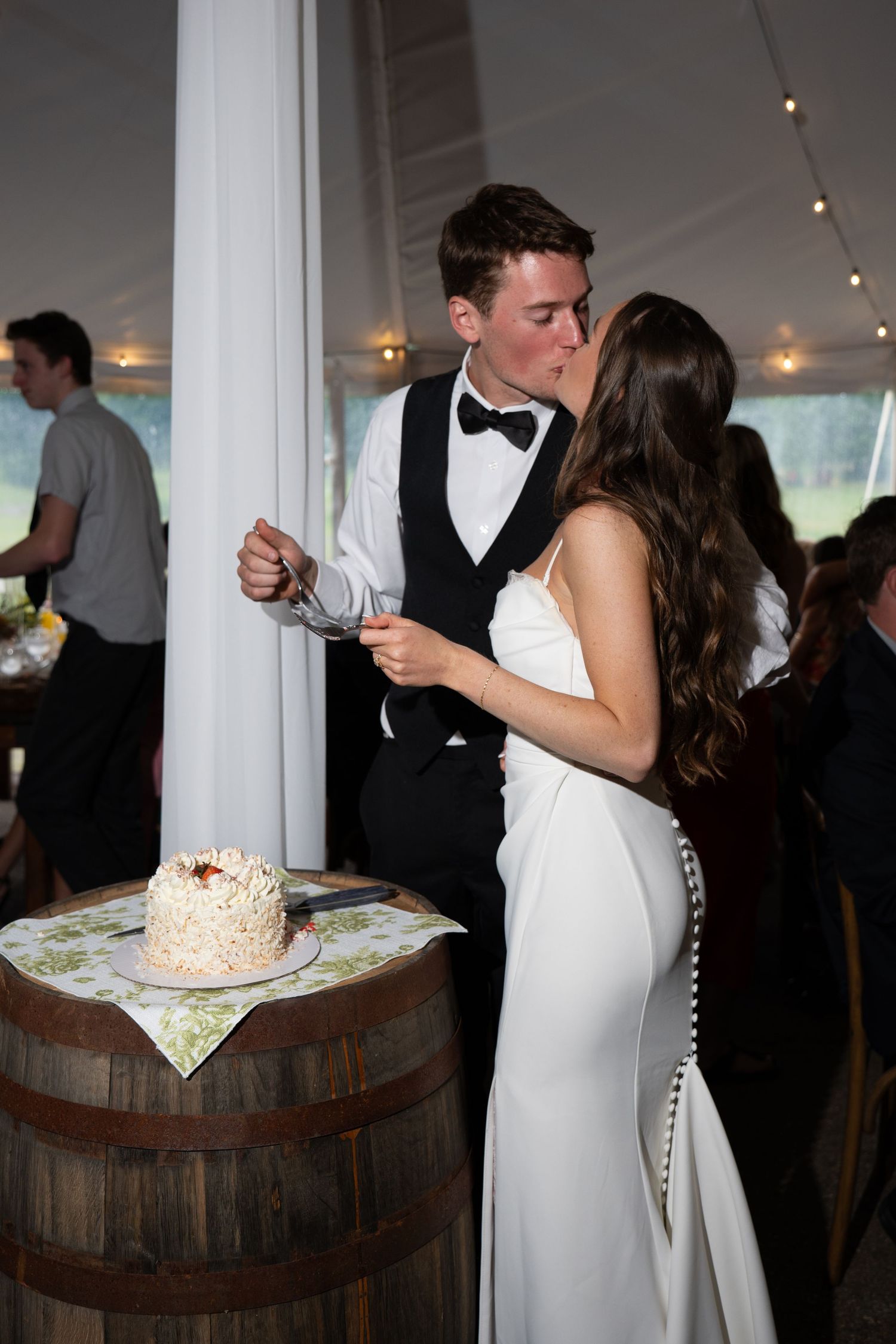 Bride and groom share a kiss while cutting their wedding cake on a barrel at an elegant tented reception.