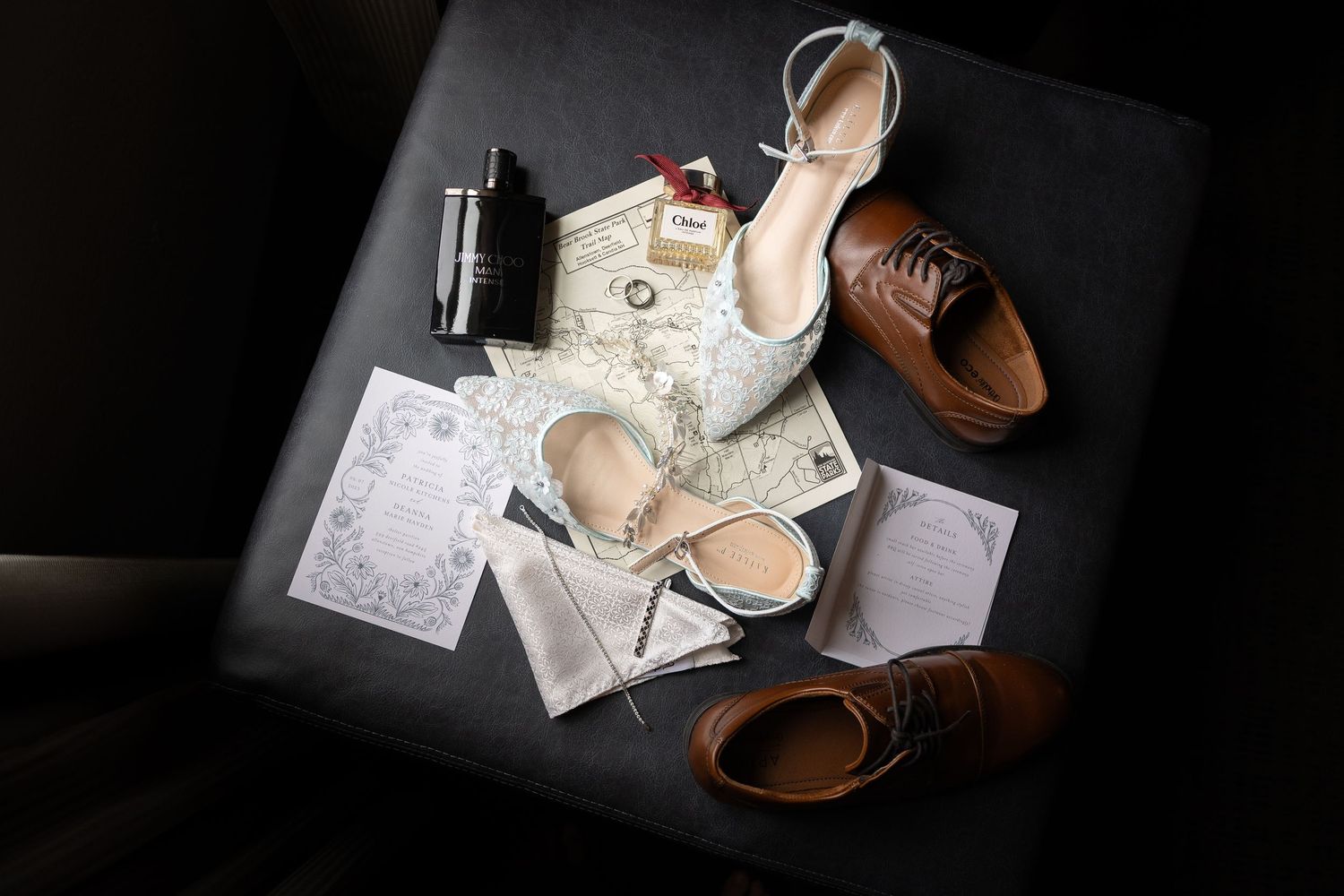 Flat lay of wedding details including brown leather shoes, white heels, map, invitations, and accessories on dark surface.