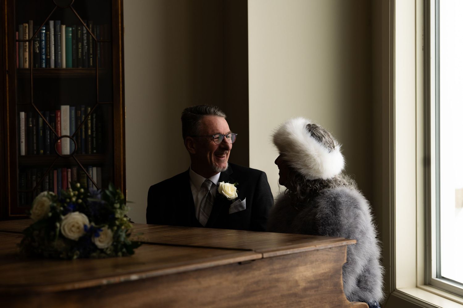 Smiling groom in tuxedo with boutonniere sitting at wooden desk with bride in winter wedding attire.