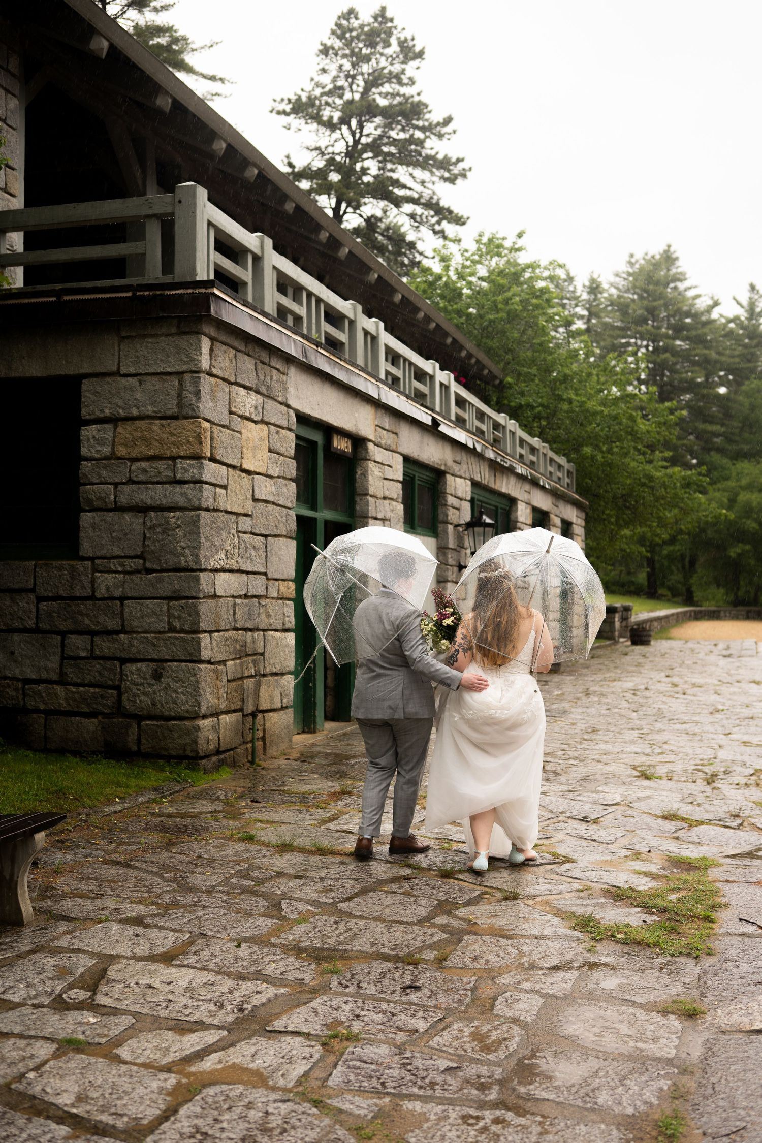Bride and groom walk with clear umbrellas in rain past a stone building.