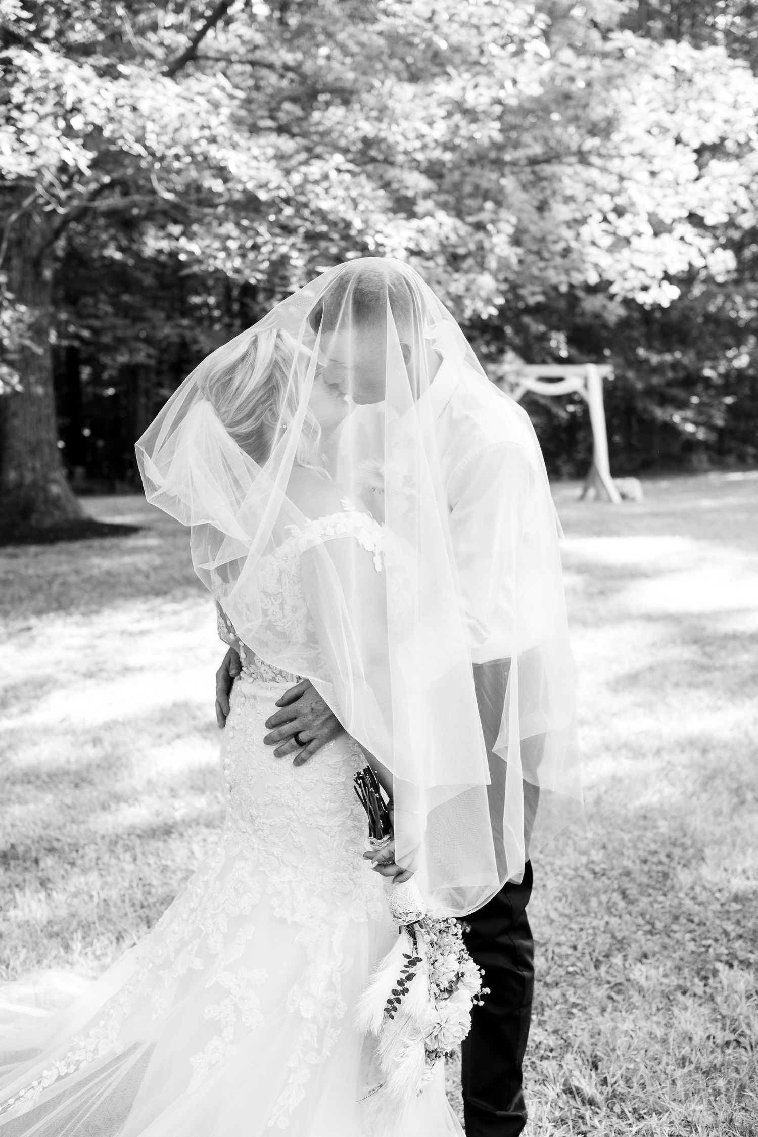 Black and white photo of bride and groom sharing intimate moment under flowing veil outdoors.