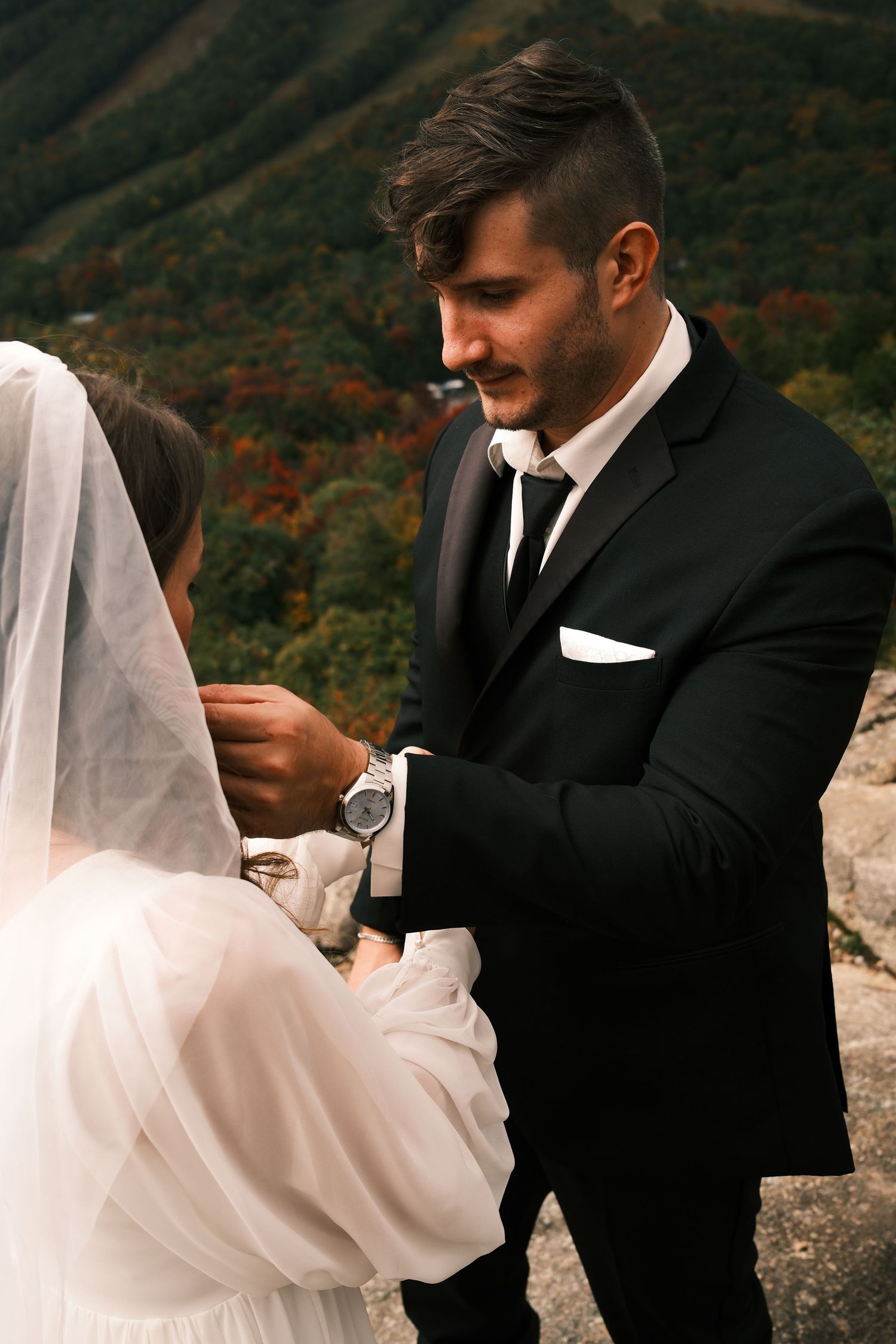 Groom in black suit adjusting brides veil with mountain landscape in background at outdoor wedding.