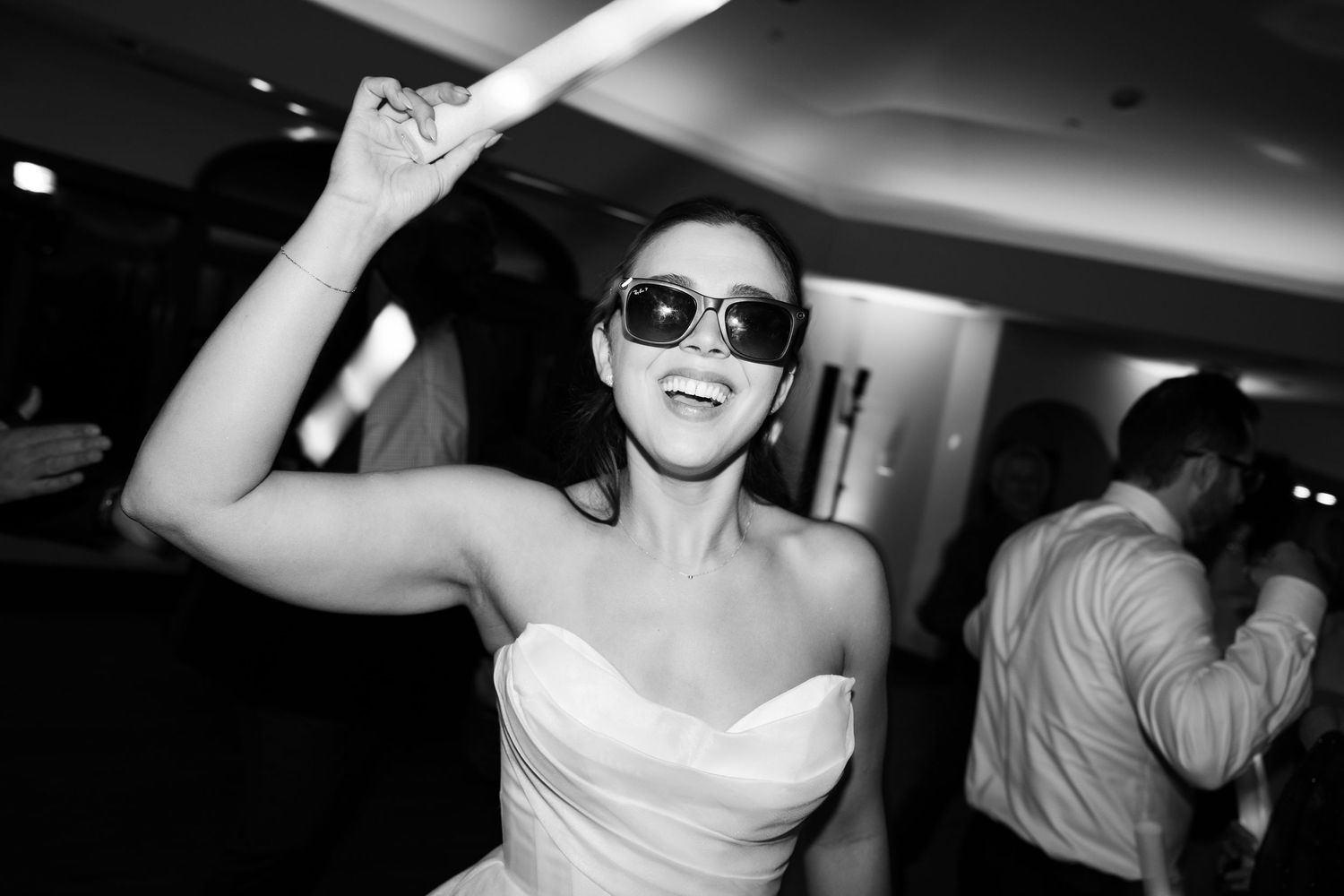 Bride in strapless white gown dancing joyfully wearing sunglasses at lively wedding reception party.