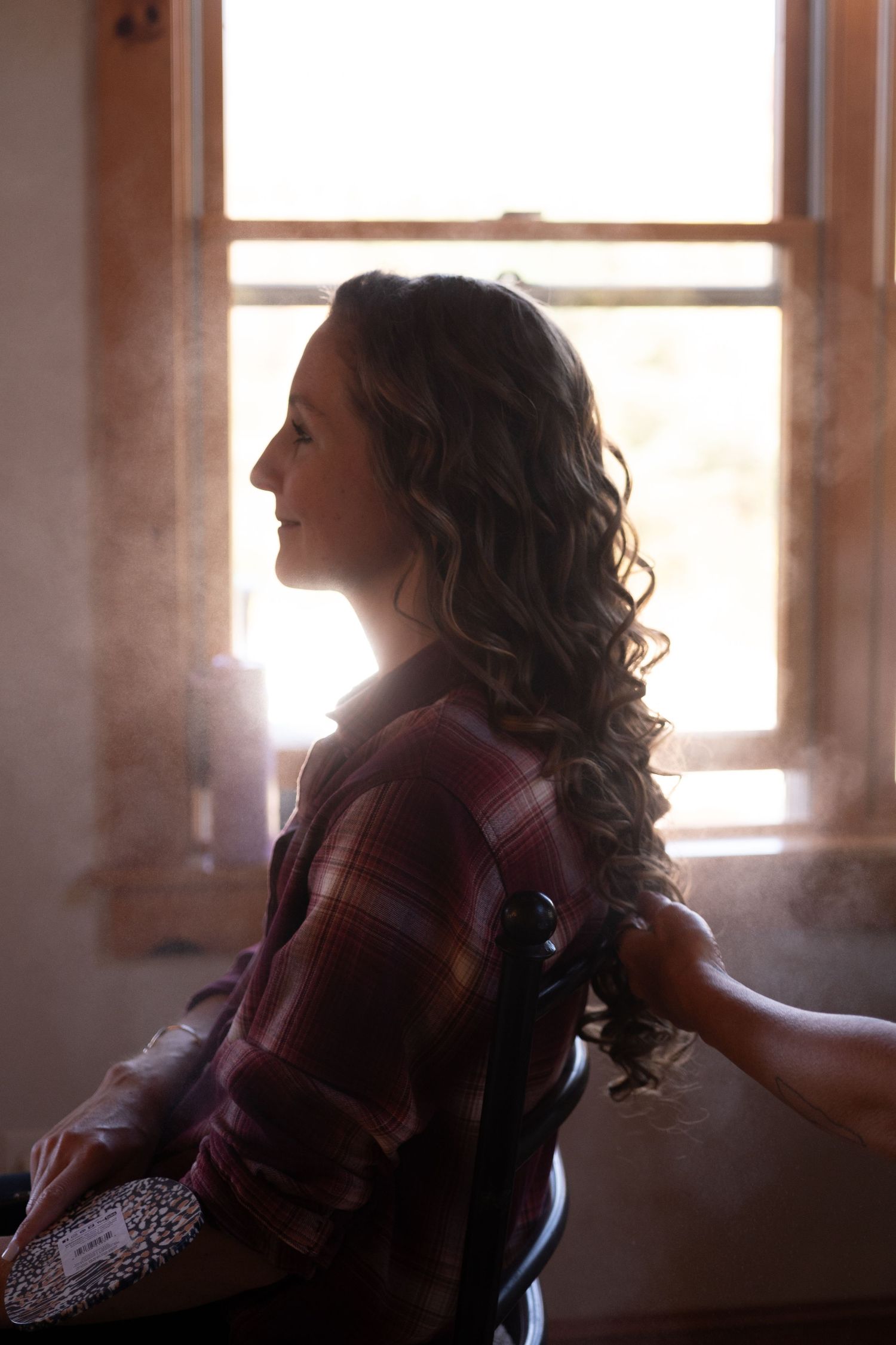 A woman with curly hair sits by a sunlit window wearing a plaid shirt, smiling softly in a warmly lit room.