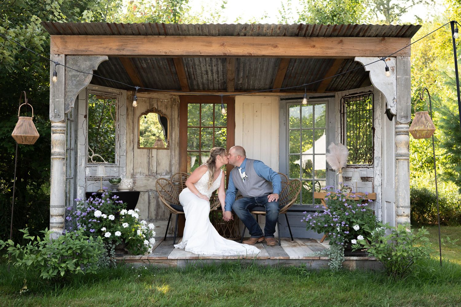 Bride and groom share a kiss under a rustic wooden gazebo decorated with string lights and flowers.