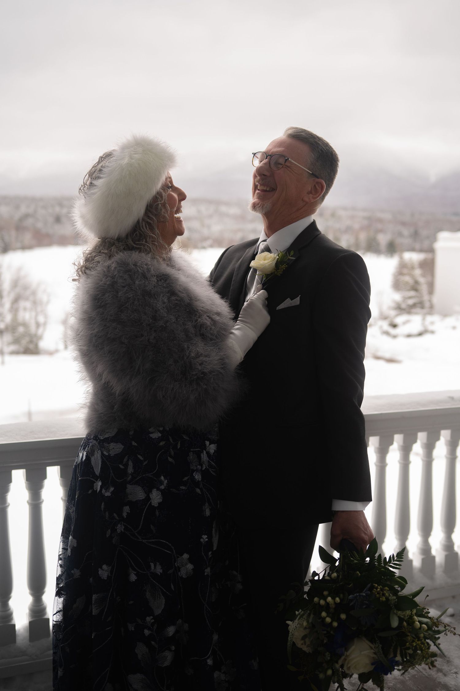Joyful mature couple in winter wedding attire laughing on snowy mountain venue balcony.