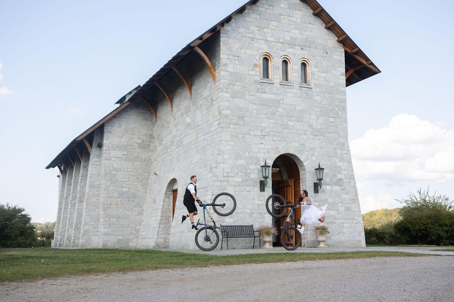 Groom performs wheelie on mountain bike outside a stone chapel as bride watches in wedding attire.