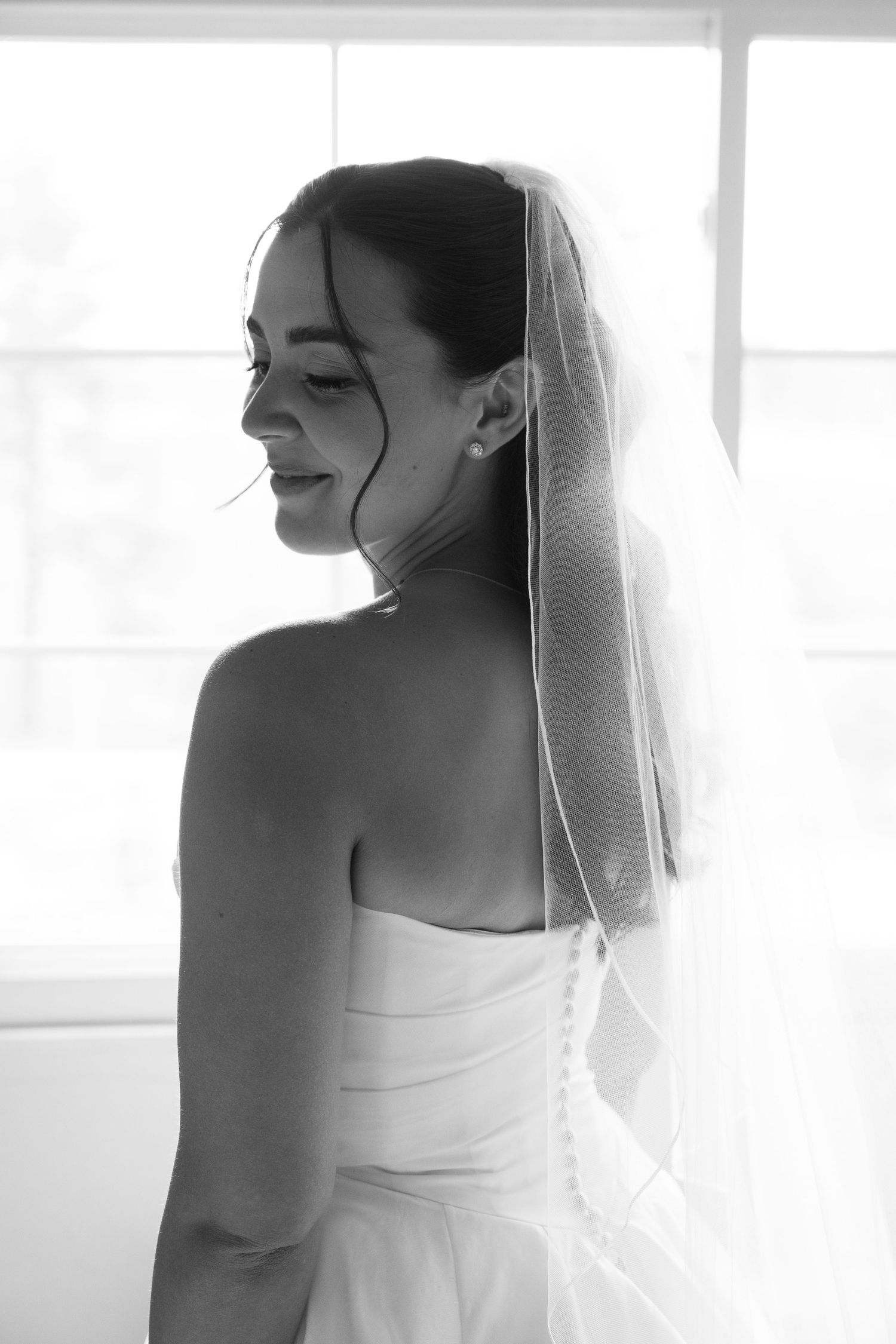 Elegant black and white portrait of smiling bride in strapless gown and veil standing by window.