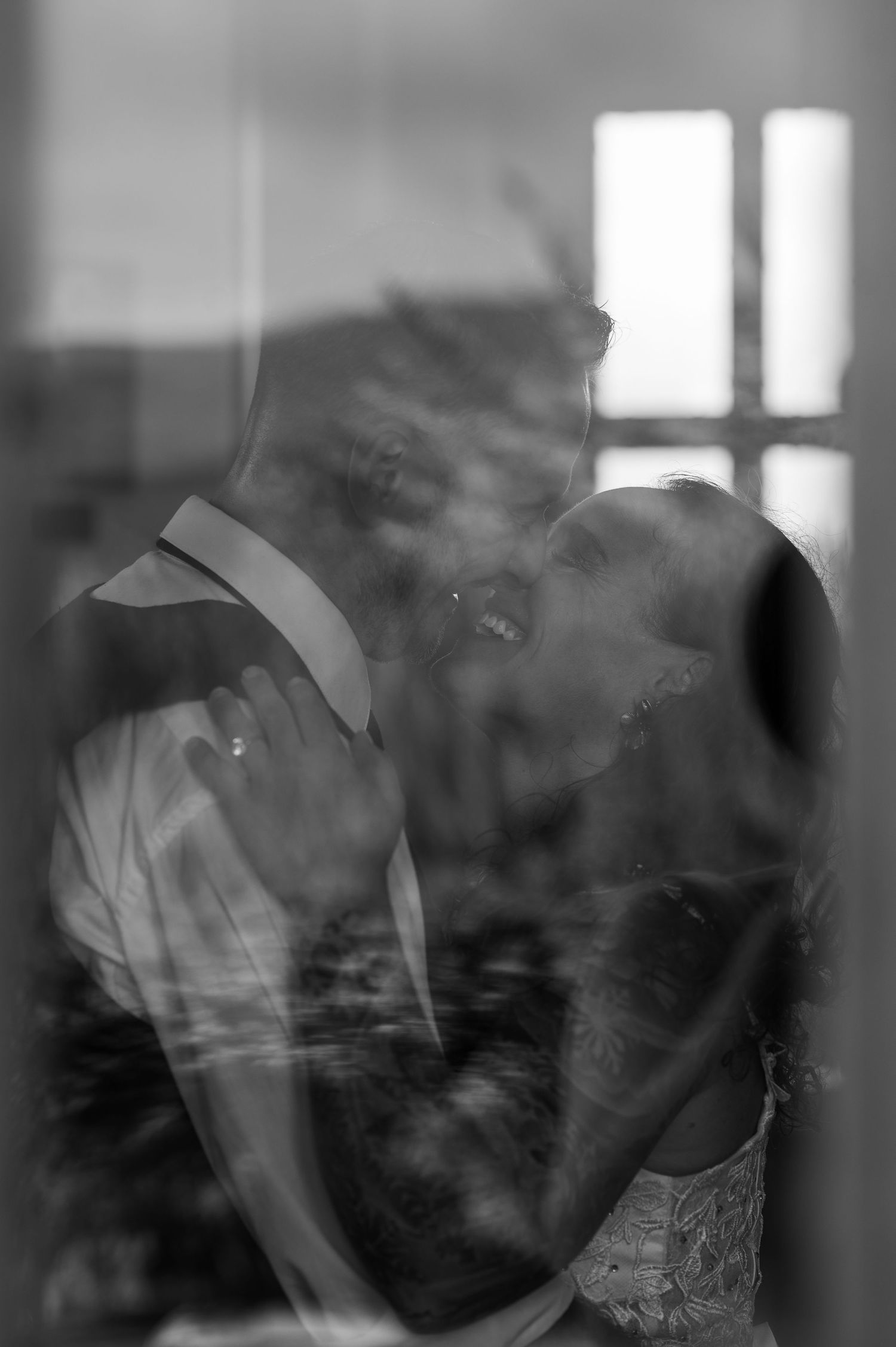 Black and white close-up of bride and groom sharing a romantic kiss through a glass window.