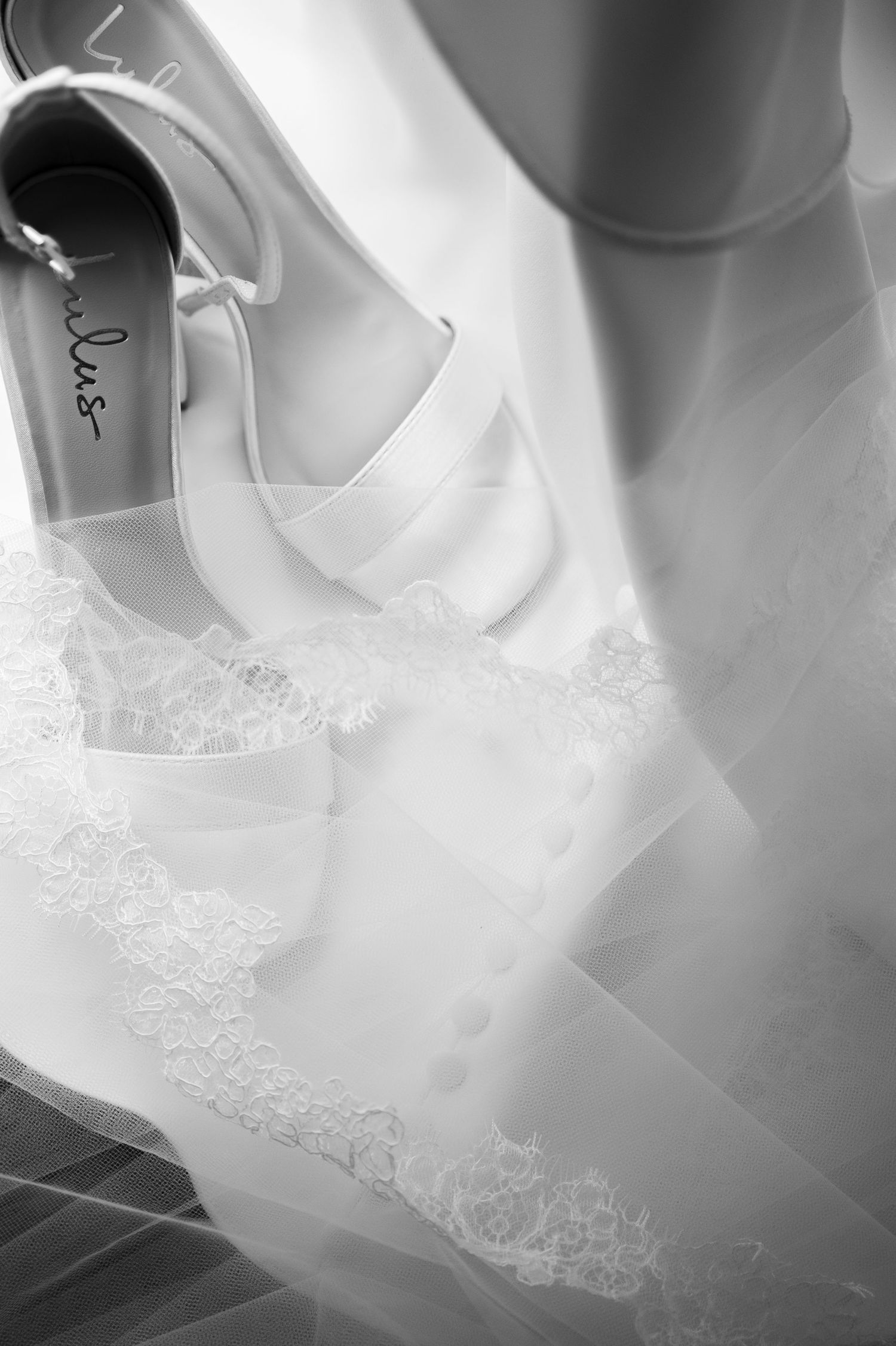 Black and white photo of elegant white bridal heels resting on delicate lace wedding veil fabric.