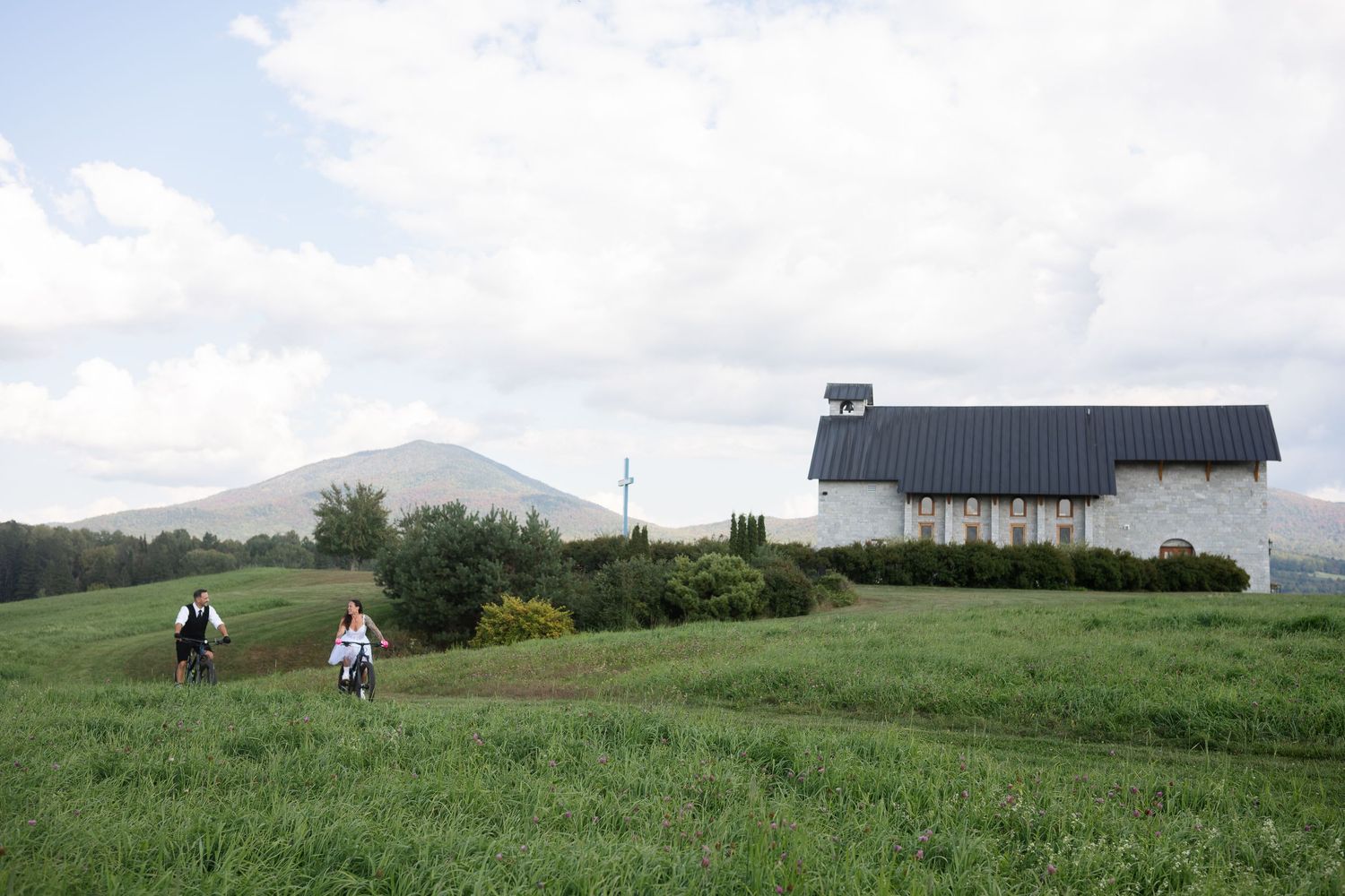 Bride and groom ride mountain bikes along a scenic green meadow path with a chapel and mountain backdrop.
