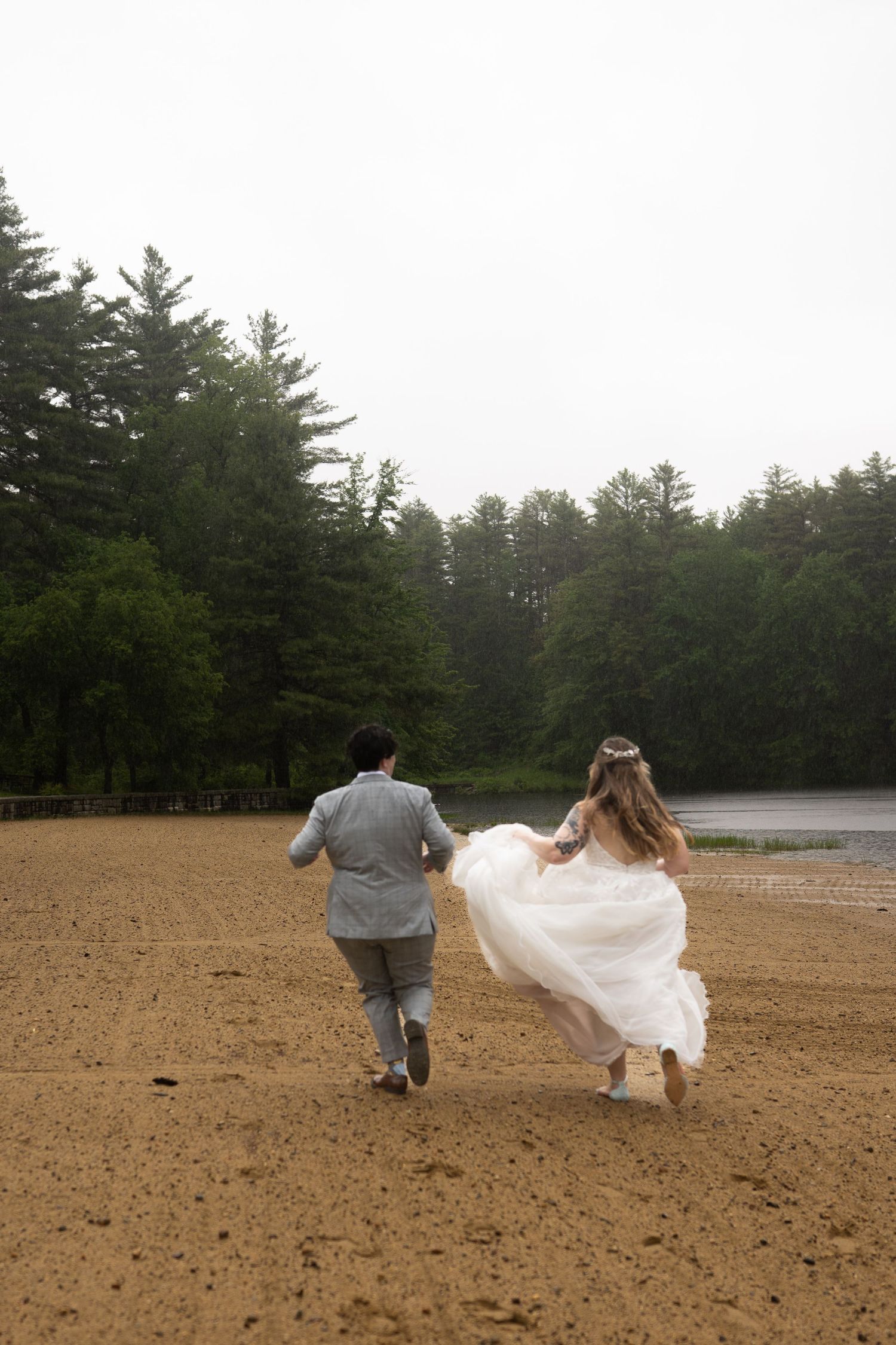 Bride in floral crown and groom run hand-in-hand across sandy beach in the rain.