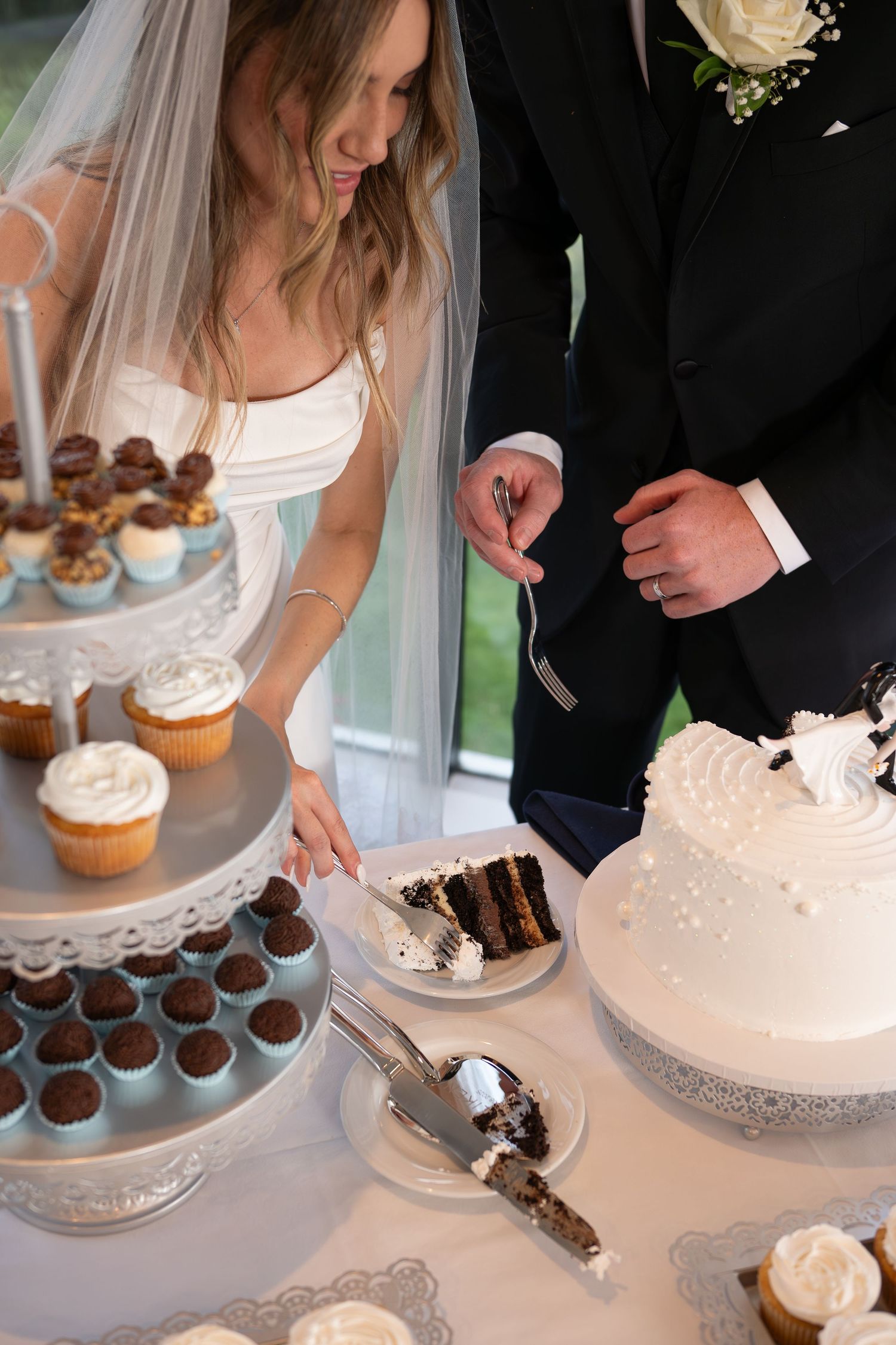 Bride and groom cut elegant white wedding cake beside tiered cupcake display at wedding reception.