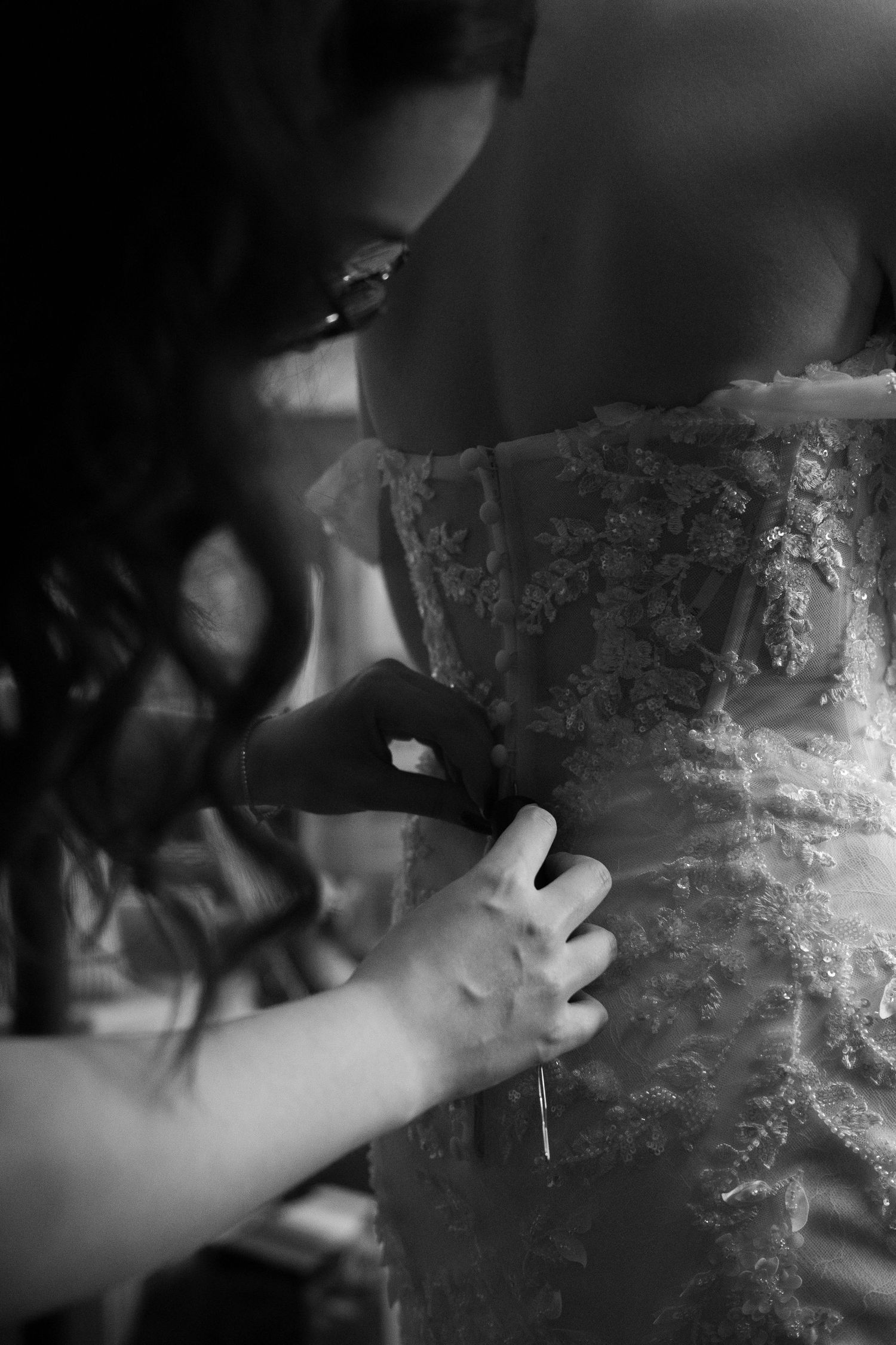 Black and white photo of bridesmaid carefully fastening buttons on brides lace wedding gown.