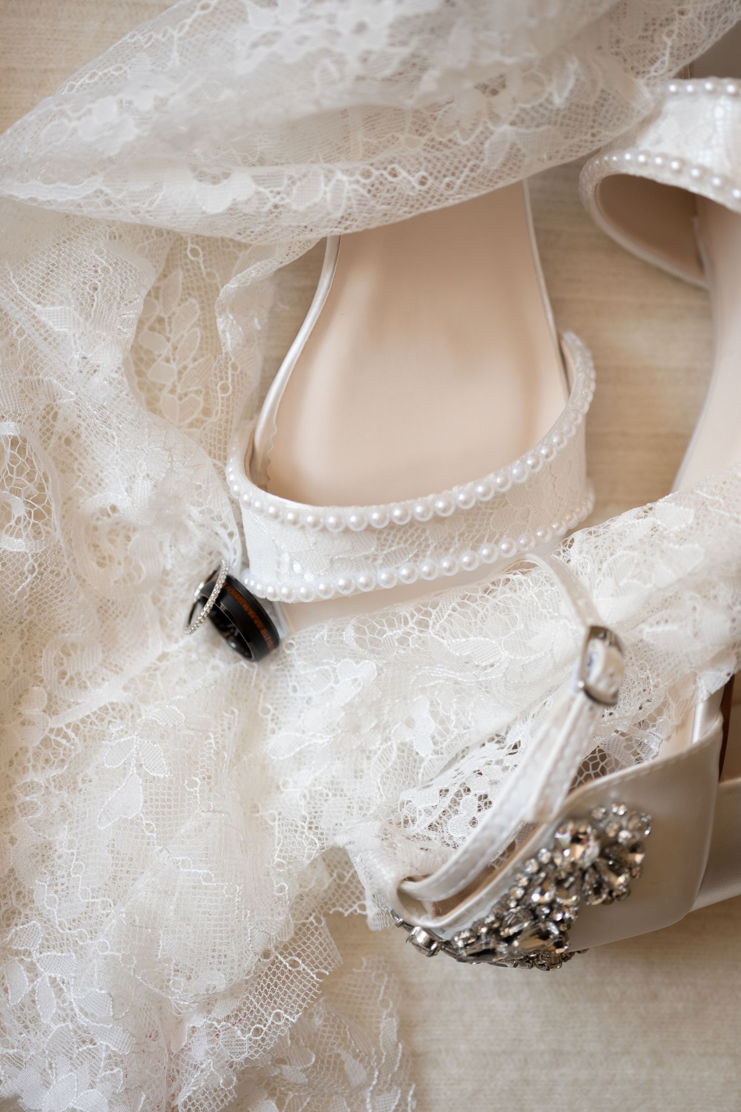 White bridal heels with pearl ankle strap and crystal embellishment resting on delicate lace wedding veil.
