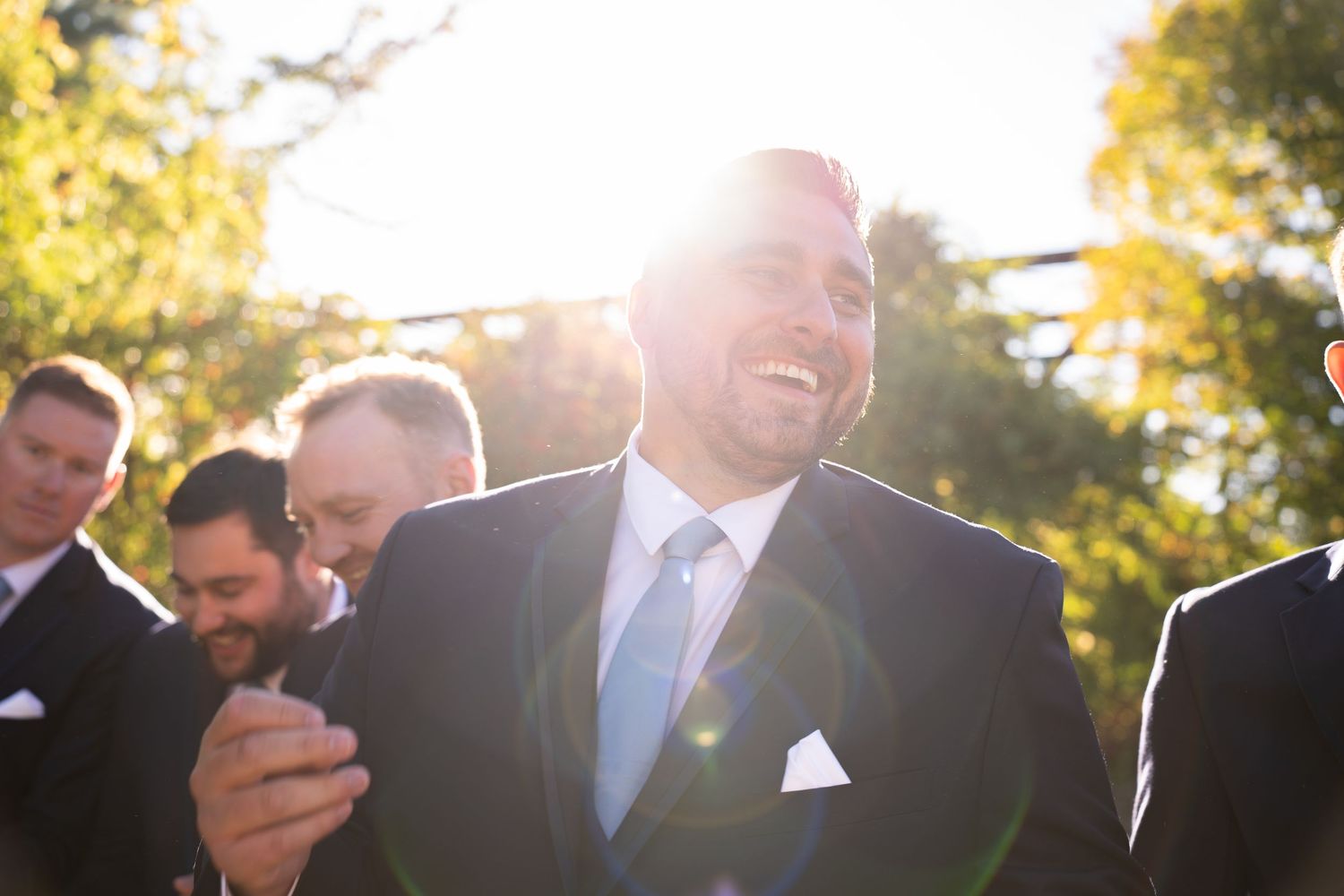 Joyful groom in navy suit laughing with groomsmen in golden sunlight during outdoor wedding celebration.
