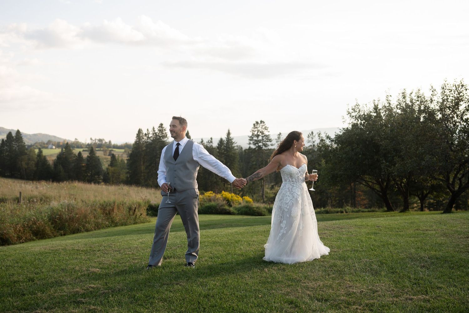 Bride and groom walking hand in hand across a green hillside at sunset.