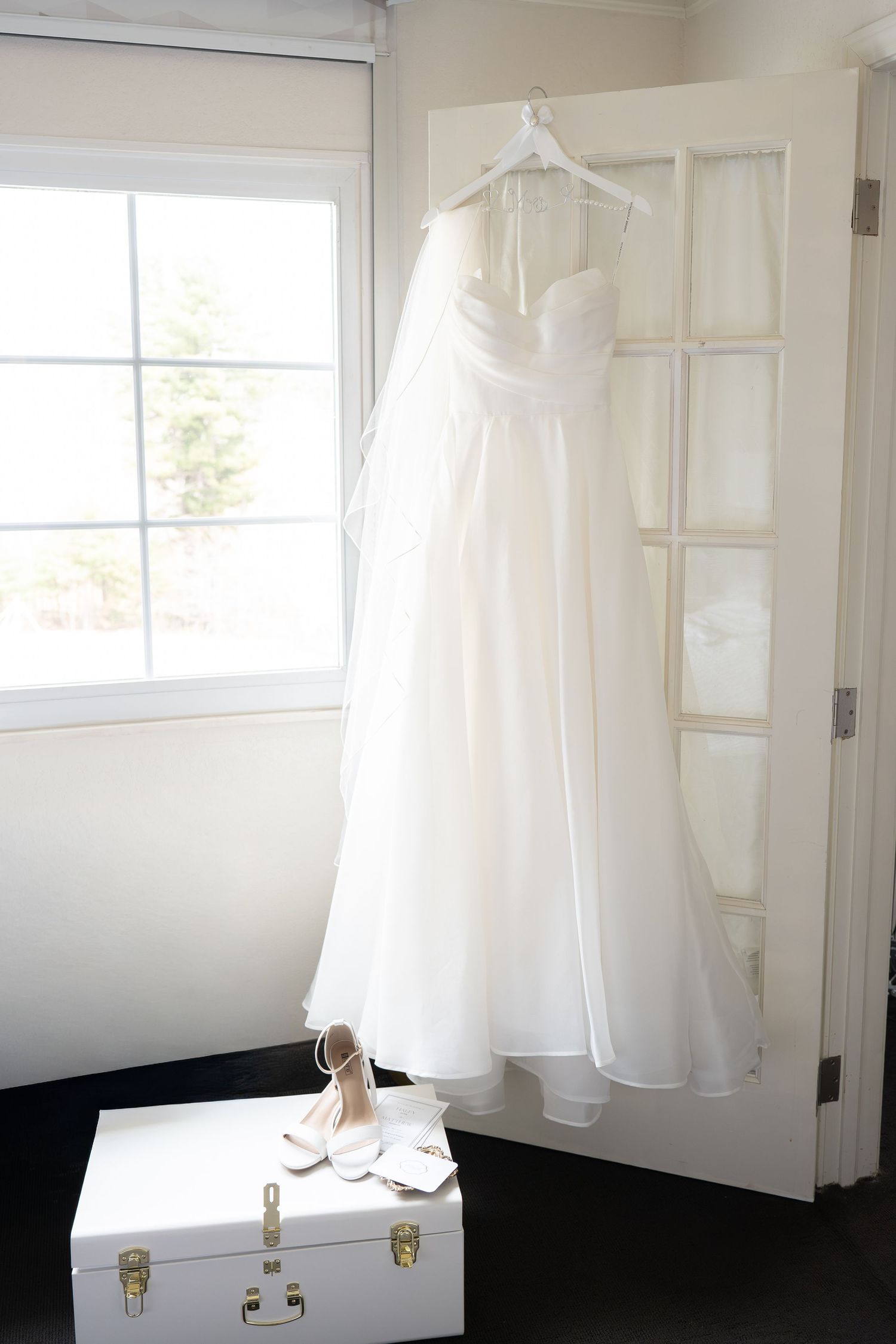 White strapless wedding gown hanging on a door beside a window with bridal heels on a trunk below.
