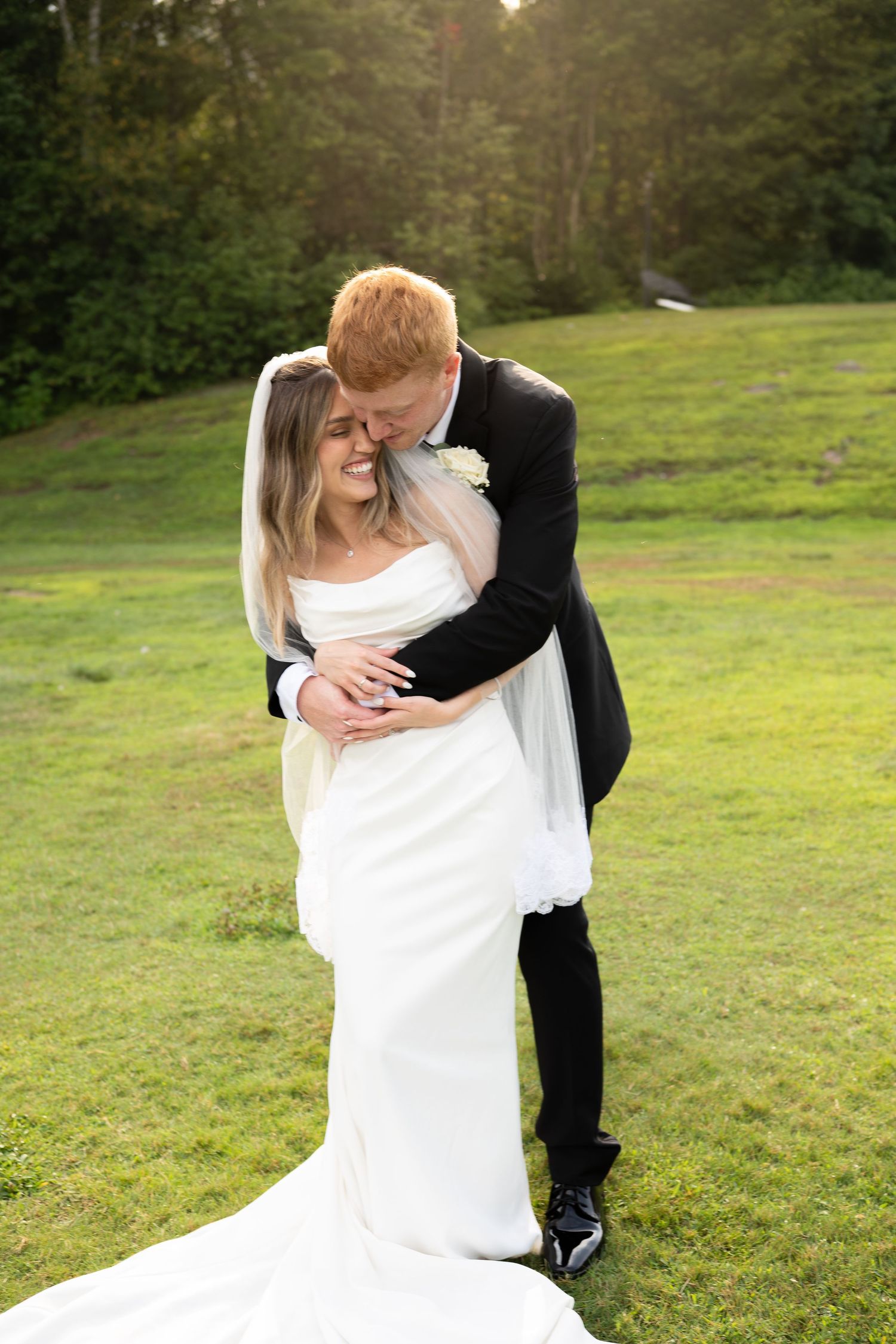 Groom in black suit embracing laughing bride in white gown on sunlit green lawn.