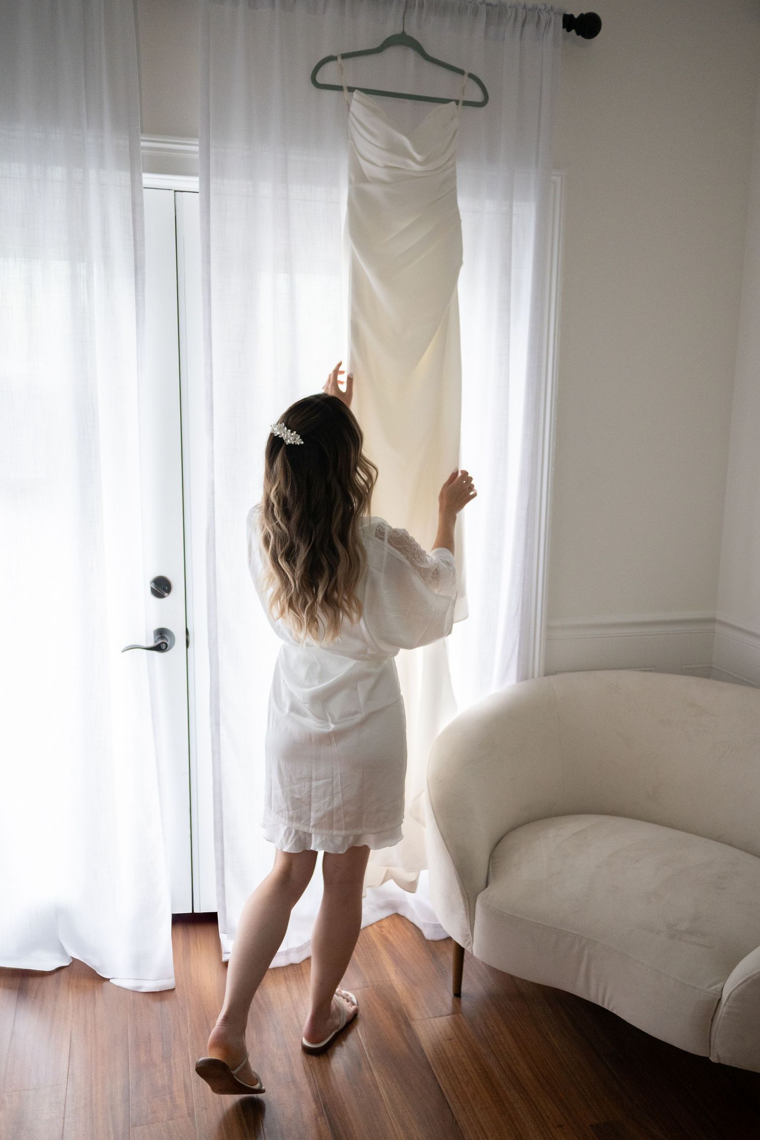 Bride in white robe reaching for wedding dress hanging by sunlit window.