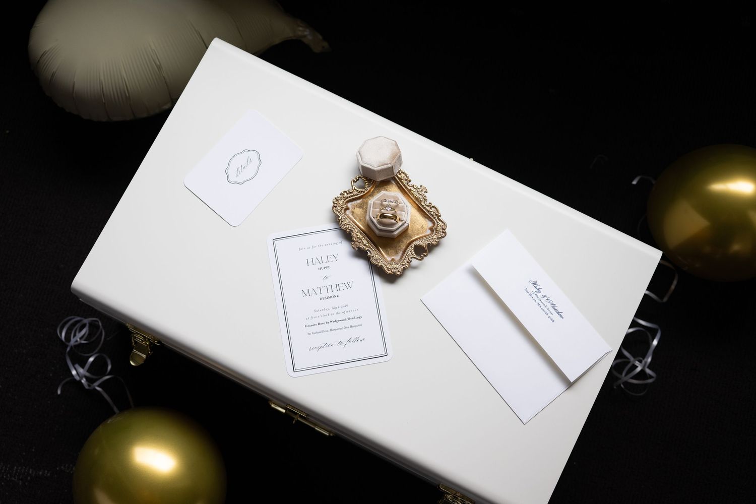 Elegant white wedding invitation box with gold ornament detail surrounded by gold and white balloons.