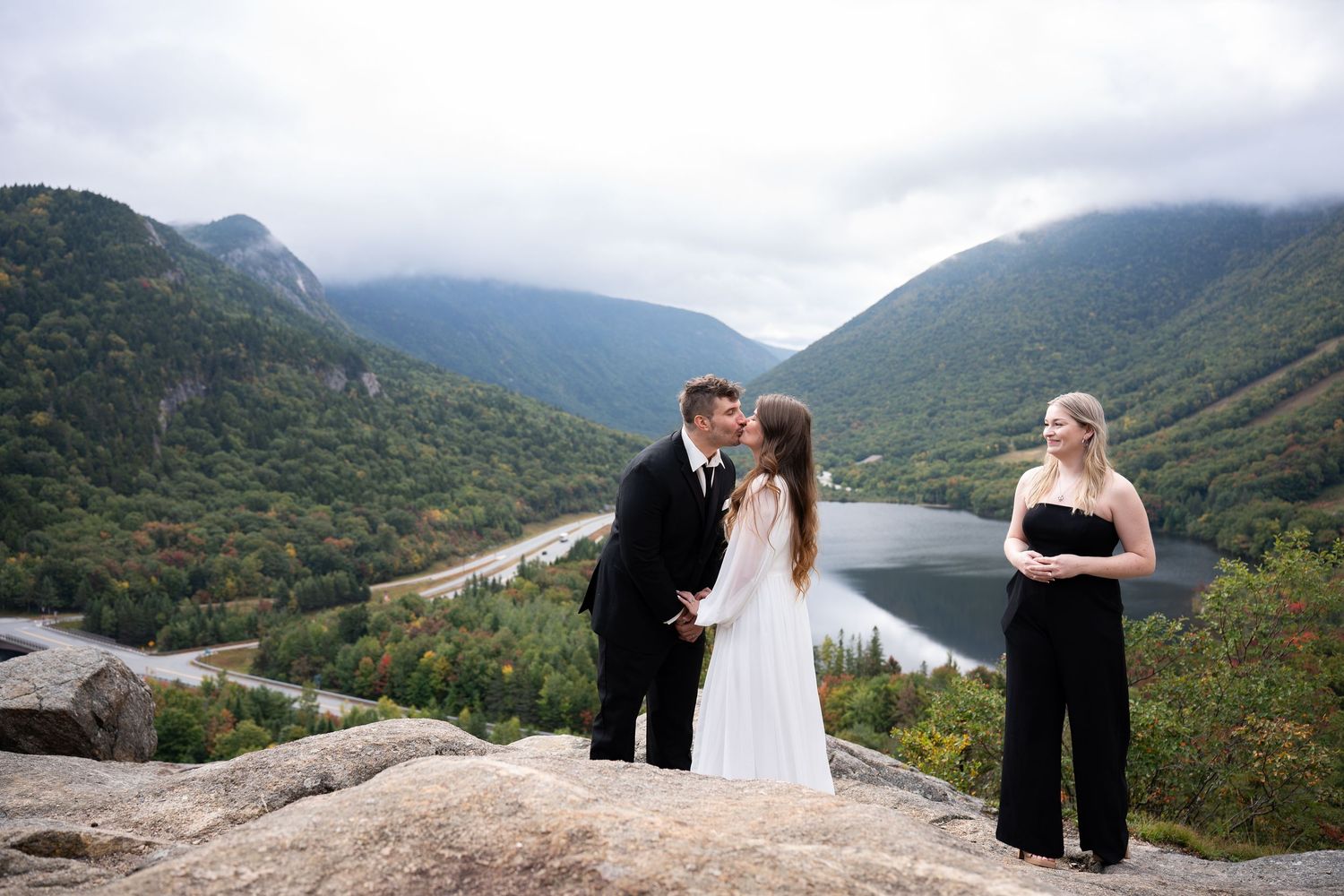 Newlyweds share first kiss on rocky mountaintop with lush green valley and lake backdrop during outdoor wedding.