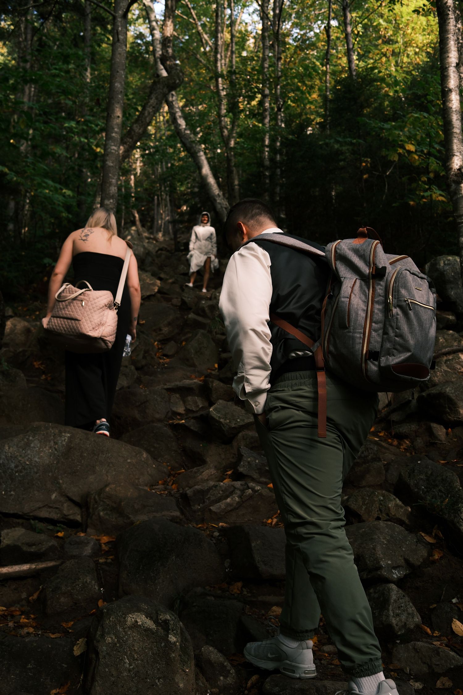 Hikers with backpacks and bags climbing rocky forest trail surrounded by lush green trees in autumn.