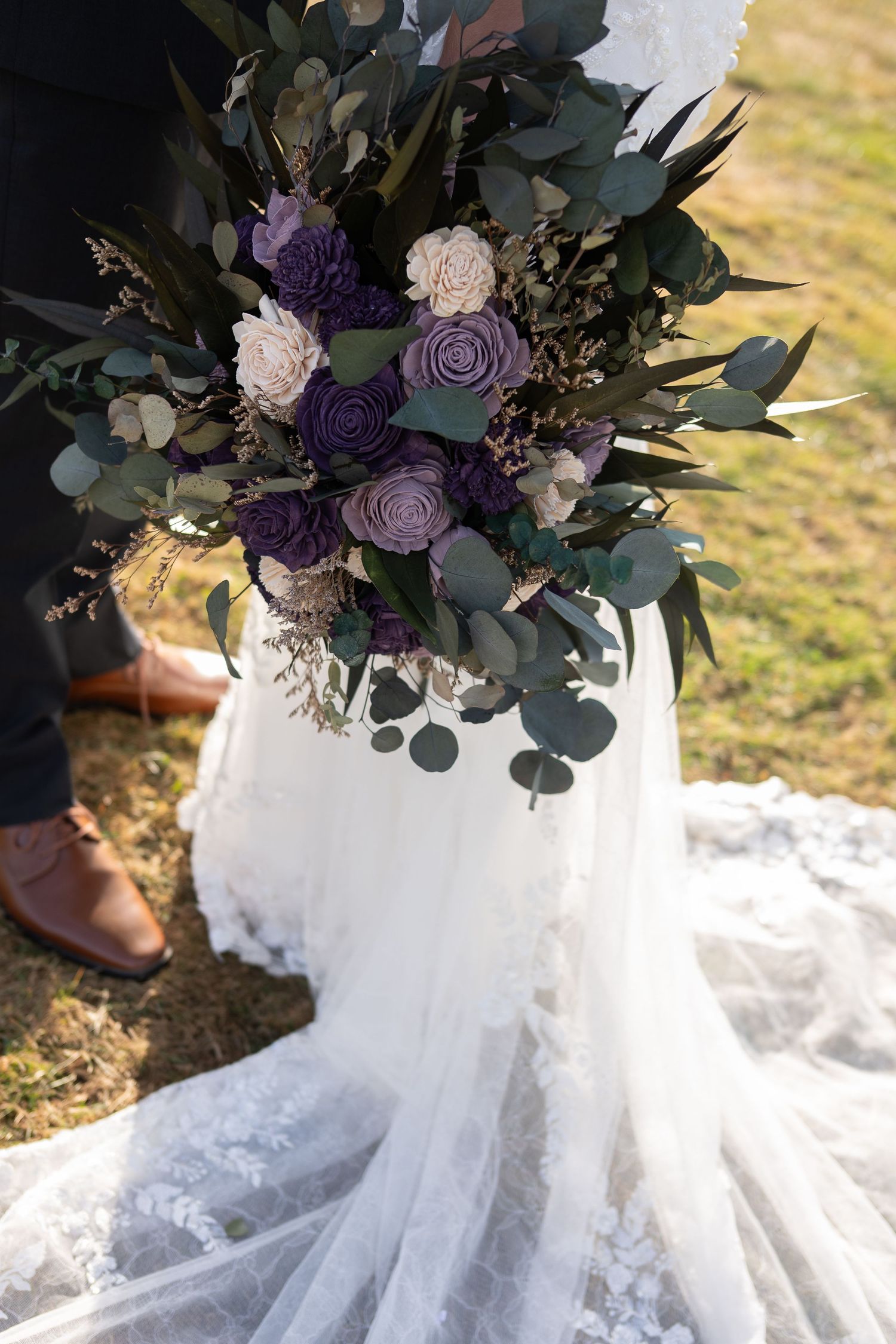 Wedding bouquet of purple and cream roses with eucalyptus held beside a lace bridal gown train.