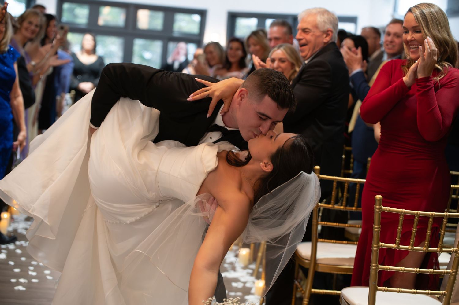 Groom dips bride for dramatic kiss during wedding reception as guests cheer and celebrate.