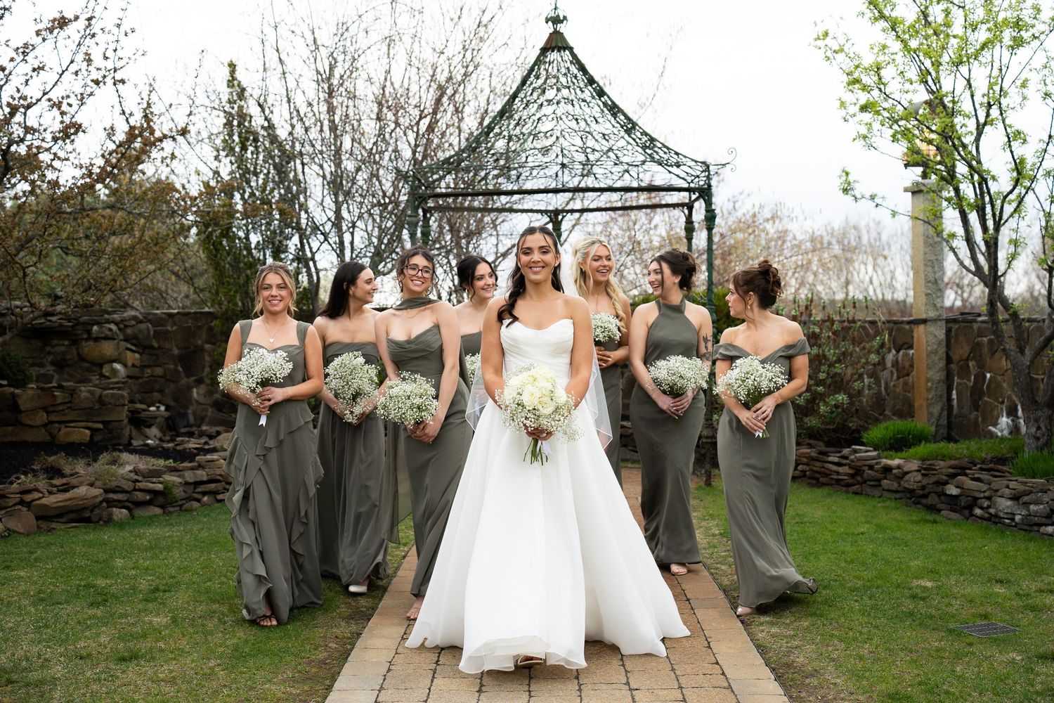 Bride in white gown leads bridesmaids in sage green dresses through garden gazebo wedding venue.
