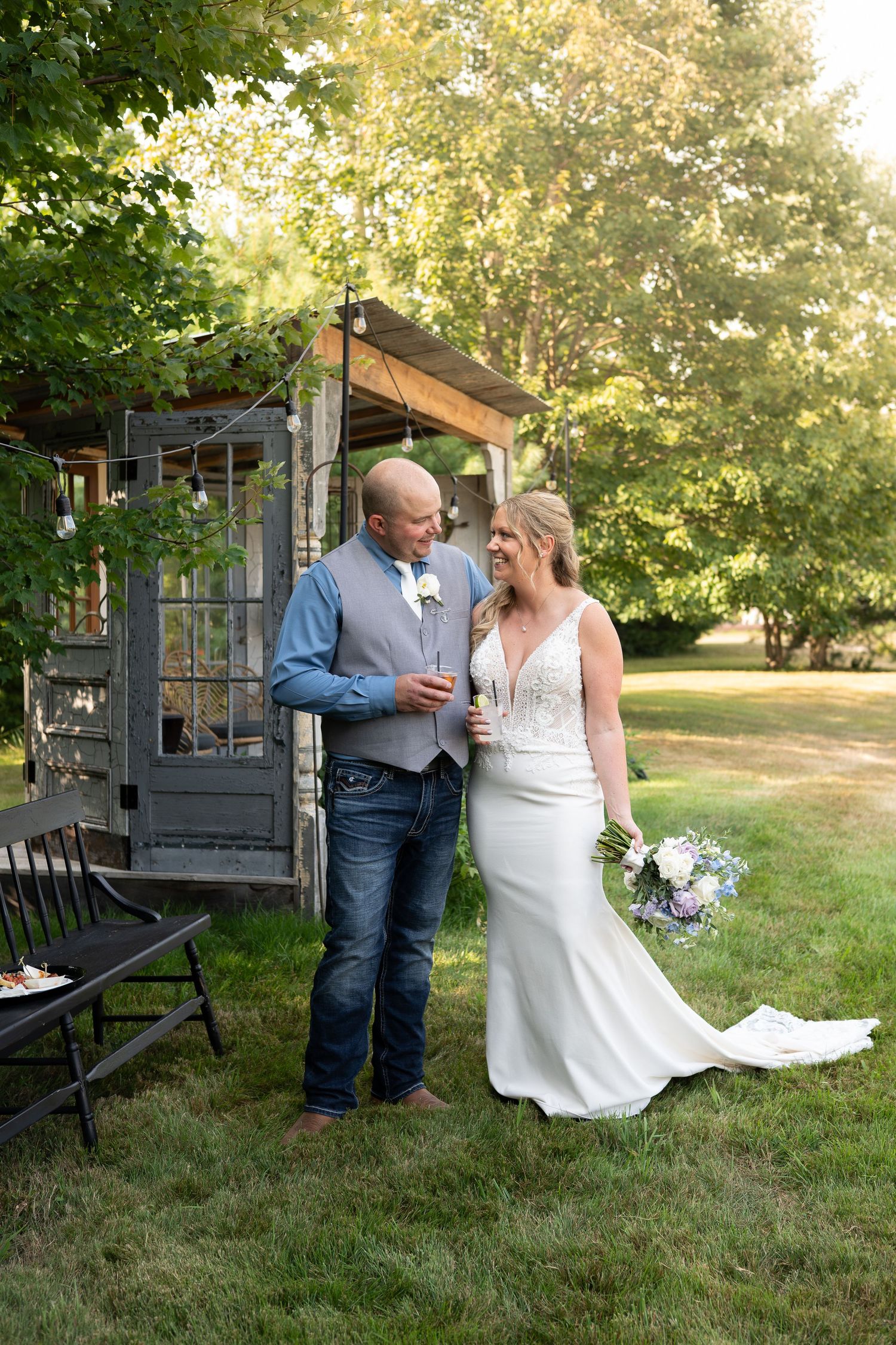 Bride holding purple bouquet and groom in blue shirt toast with champagne beside rustic wooden structure.
