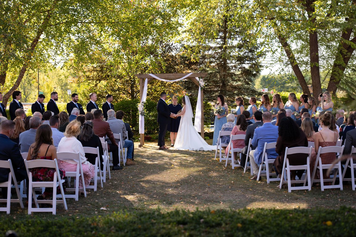 Outdoor wedding ceremony with guests seated in white chairs beneath tall trees as couple exchanges vows at altar.