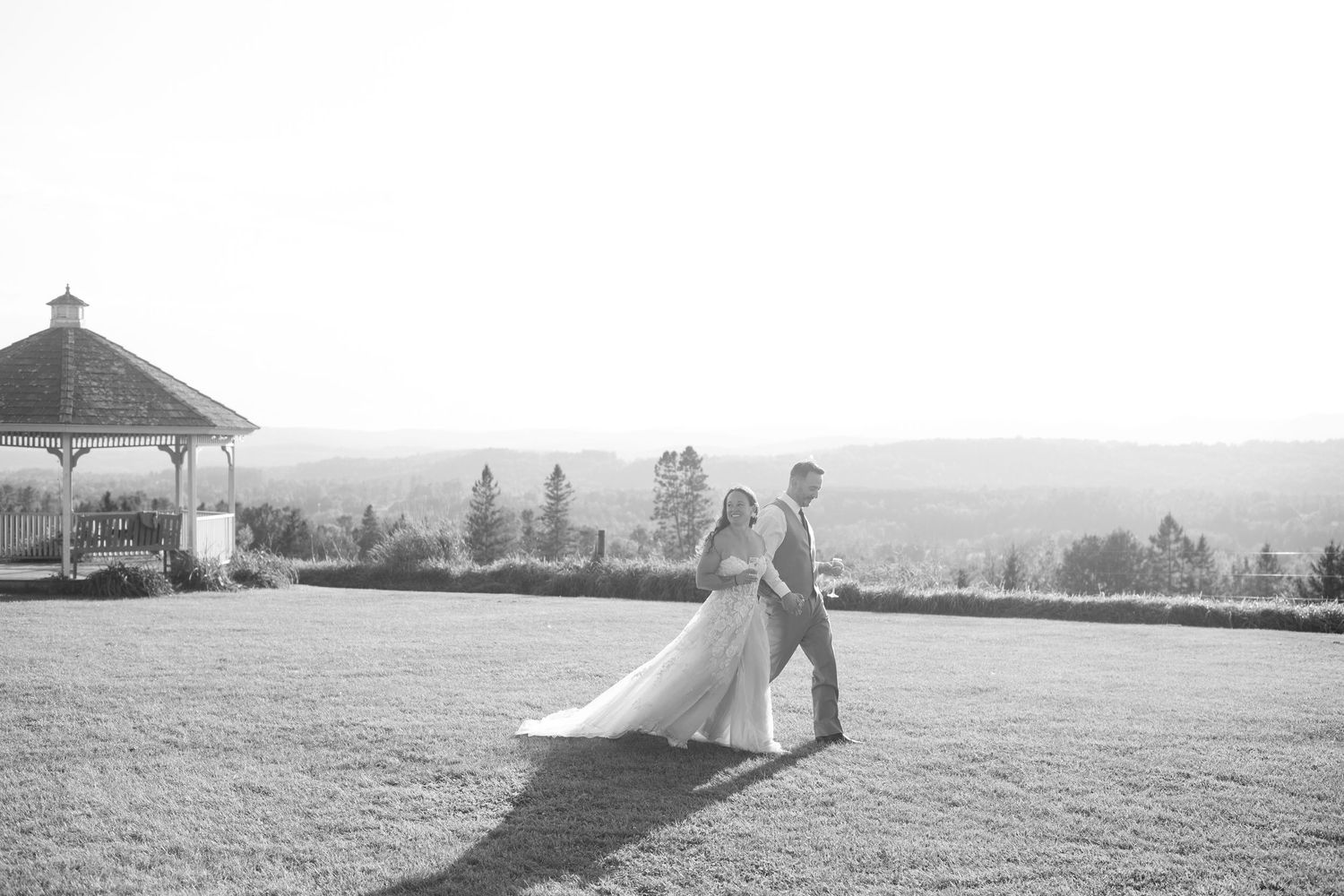 Black and white photo of bride and groom walking hand in hand near a gazebo.