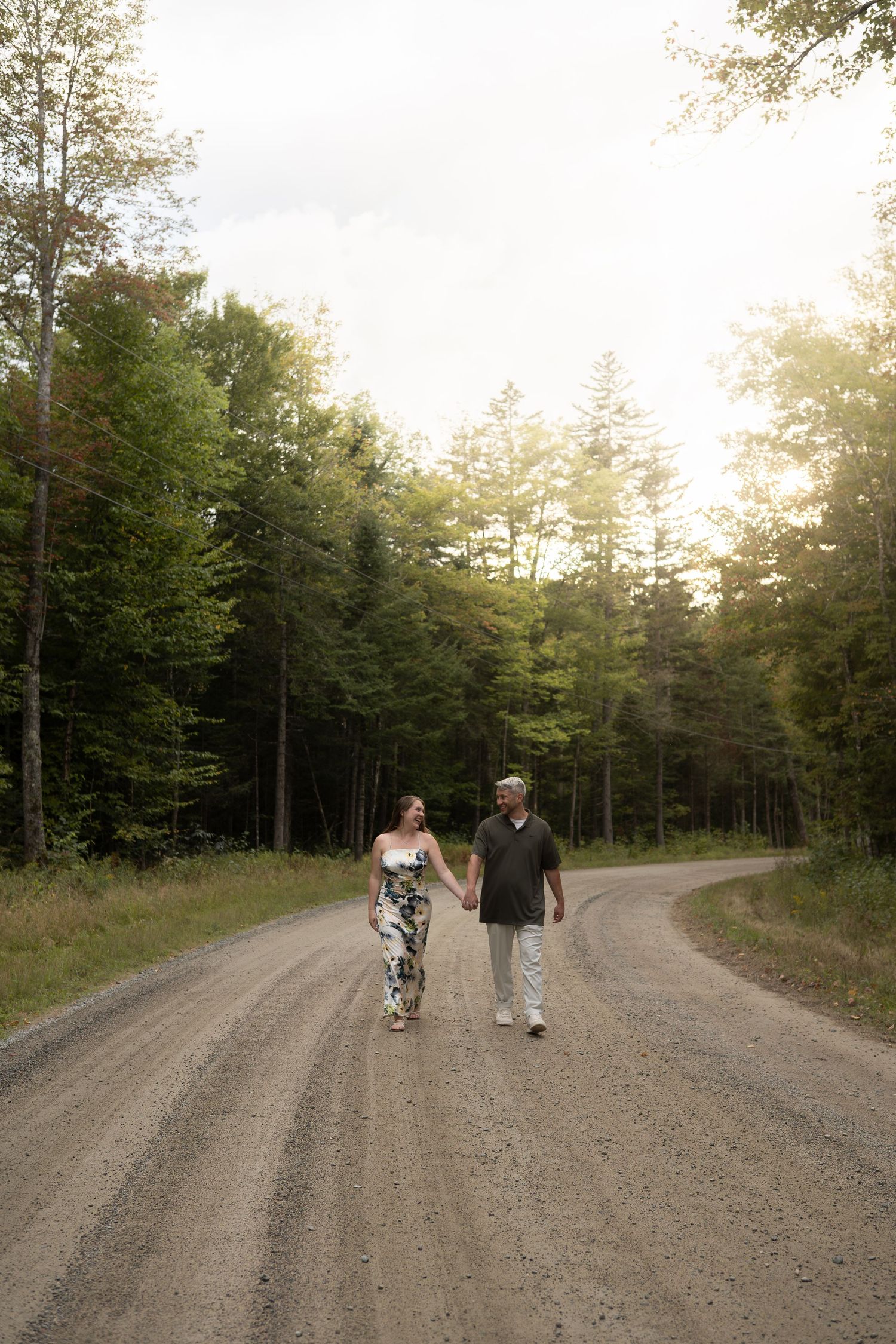 Couple walking hand in hand down a sunlit dirt road surrounded by tall trees.