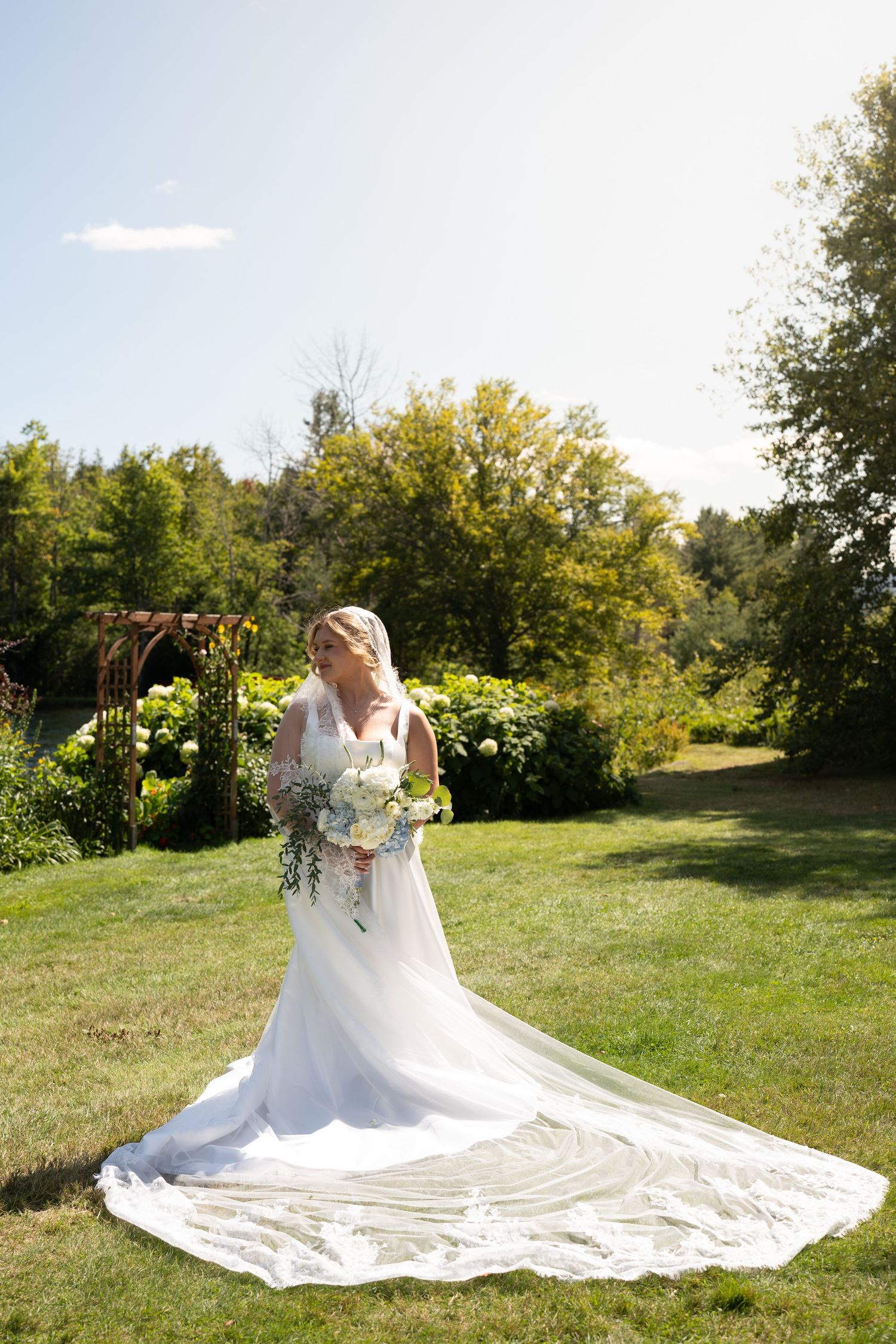 Bride in a white gown with long train holds a bouquet in a sunny garden setting.