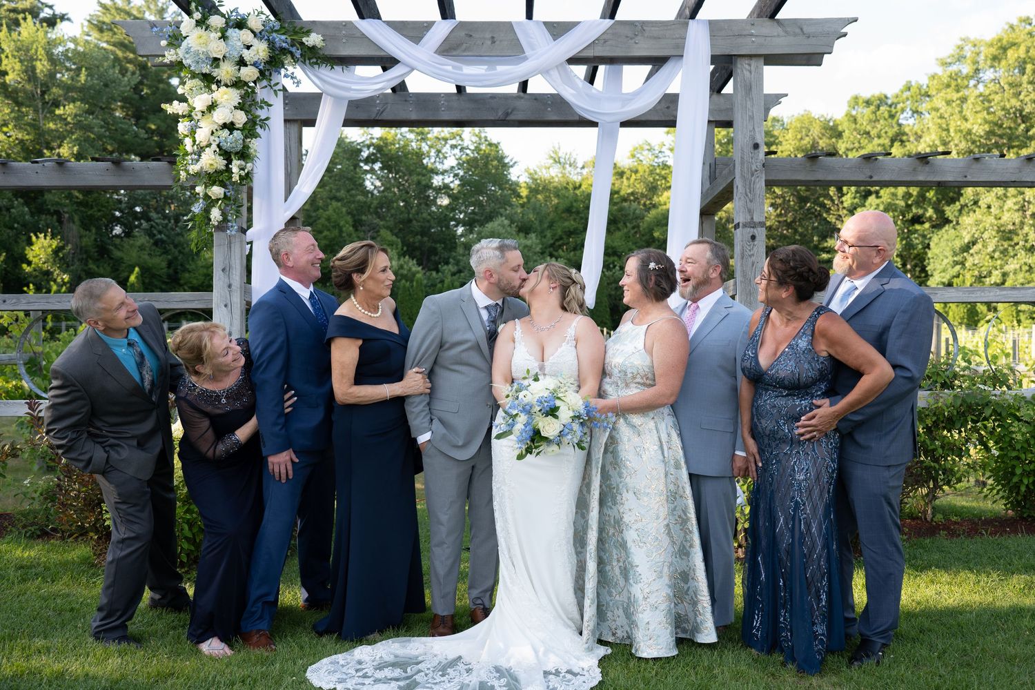 Two brides kiss surrounded by wedding party in navy attire under a white floral pergola outdoors.