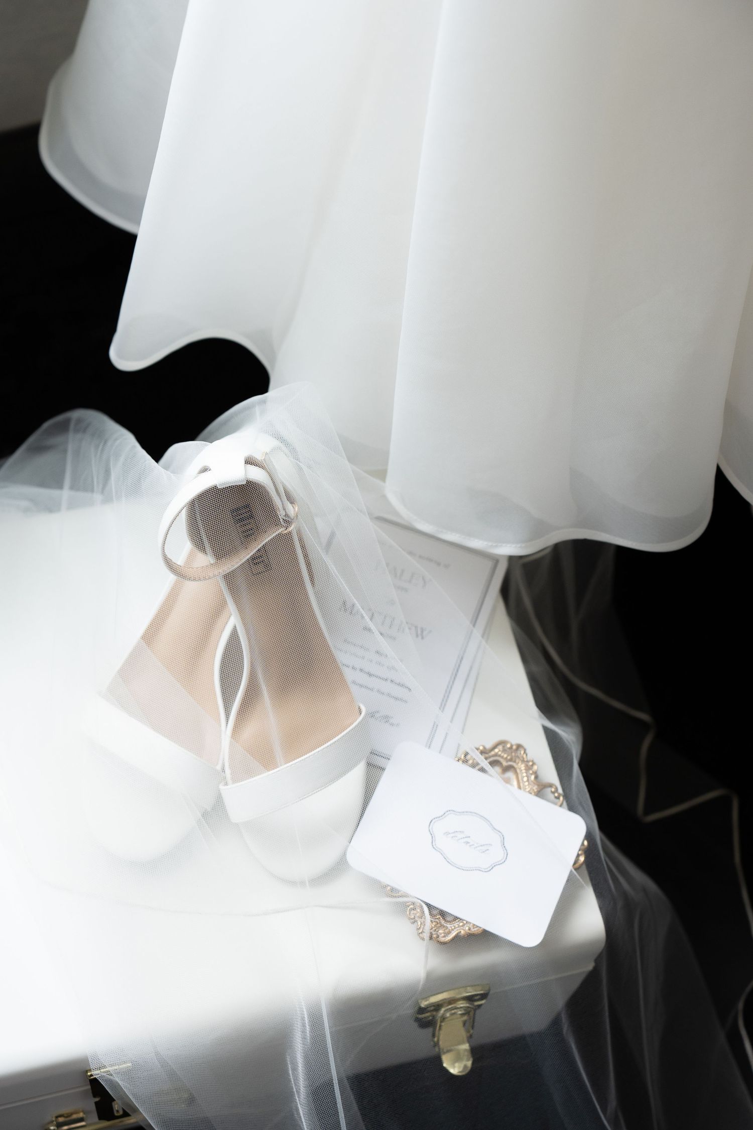 White block heel bridal shoes and small clutch purse arranged beneath flowing wedding veil on white surface.