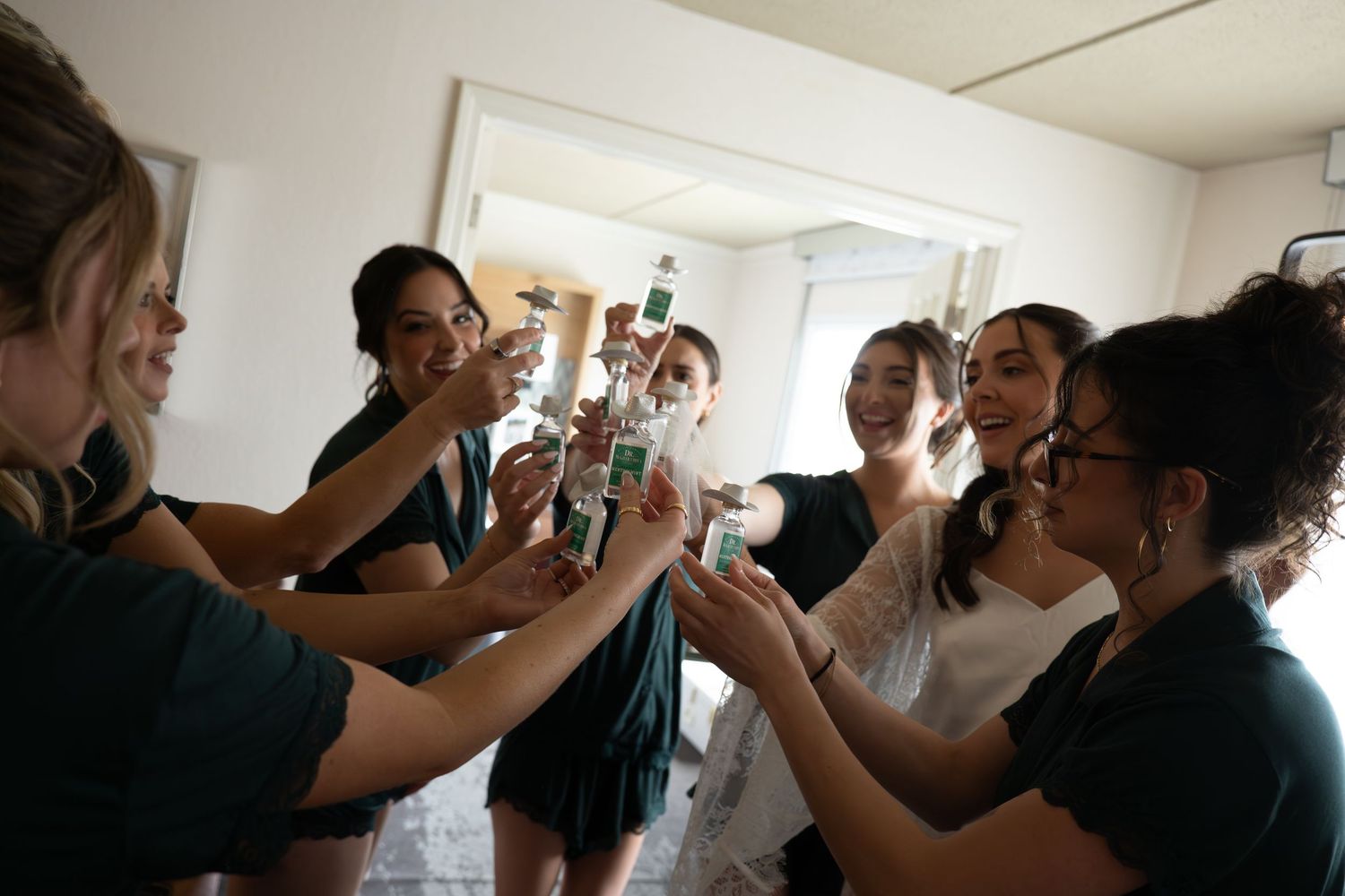 Bridesmaids in dark green dresses toasting with bottles alongside bride in white wedding dress indoors.