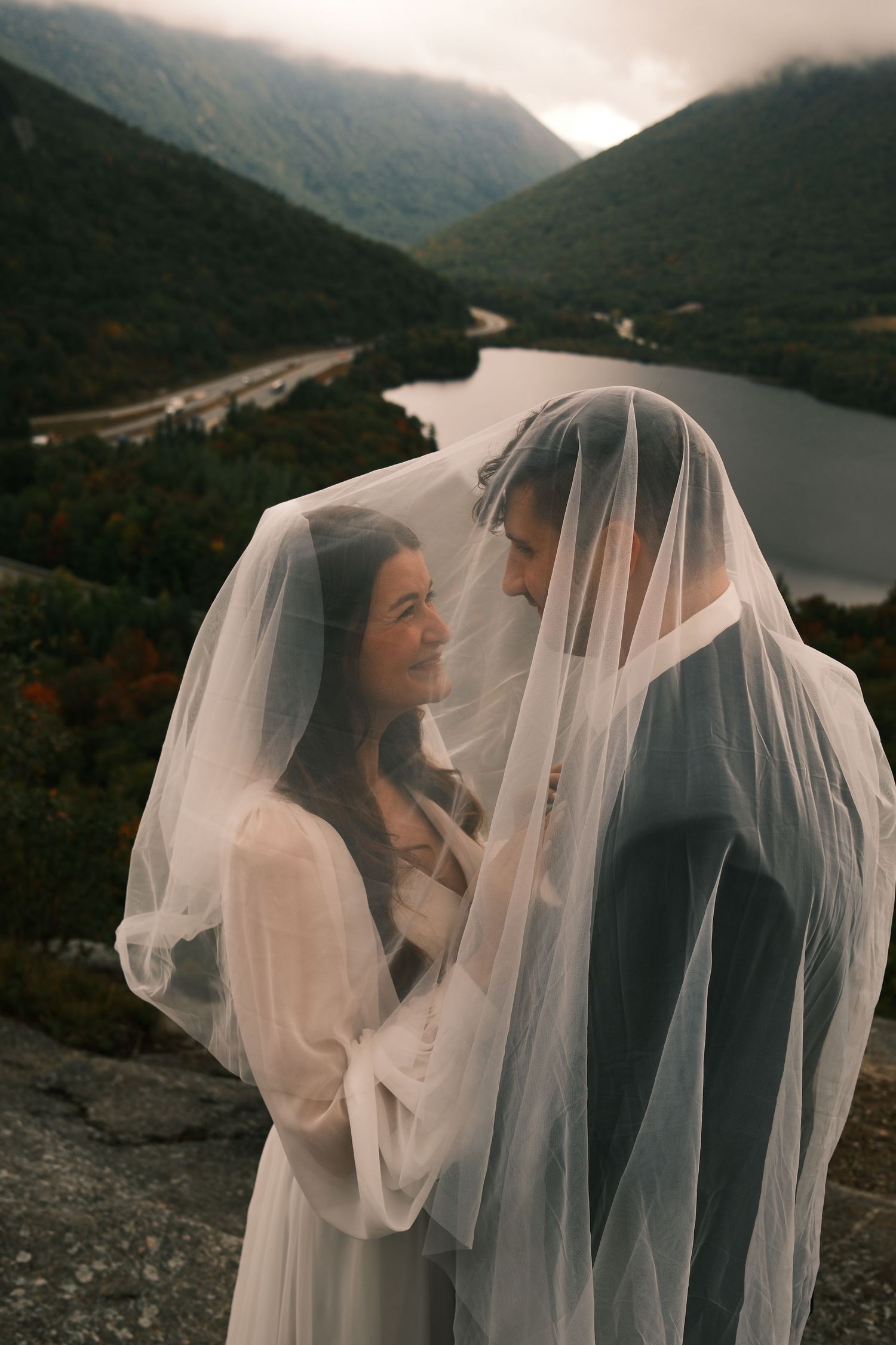 Bride and groom share romantic moment under wedding veil with mountain lake backdrop during outdoor ceremony.