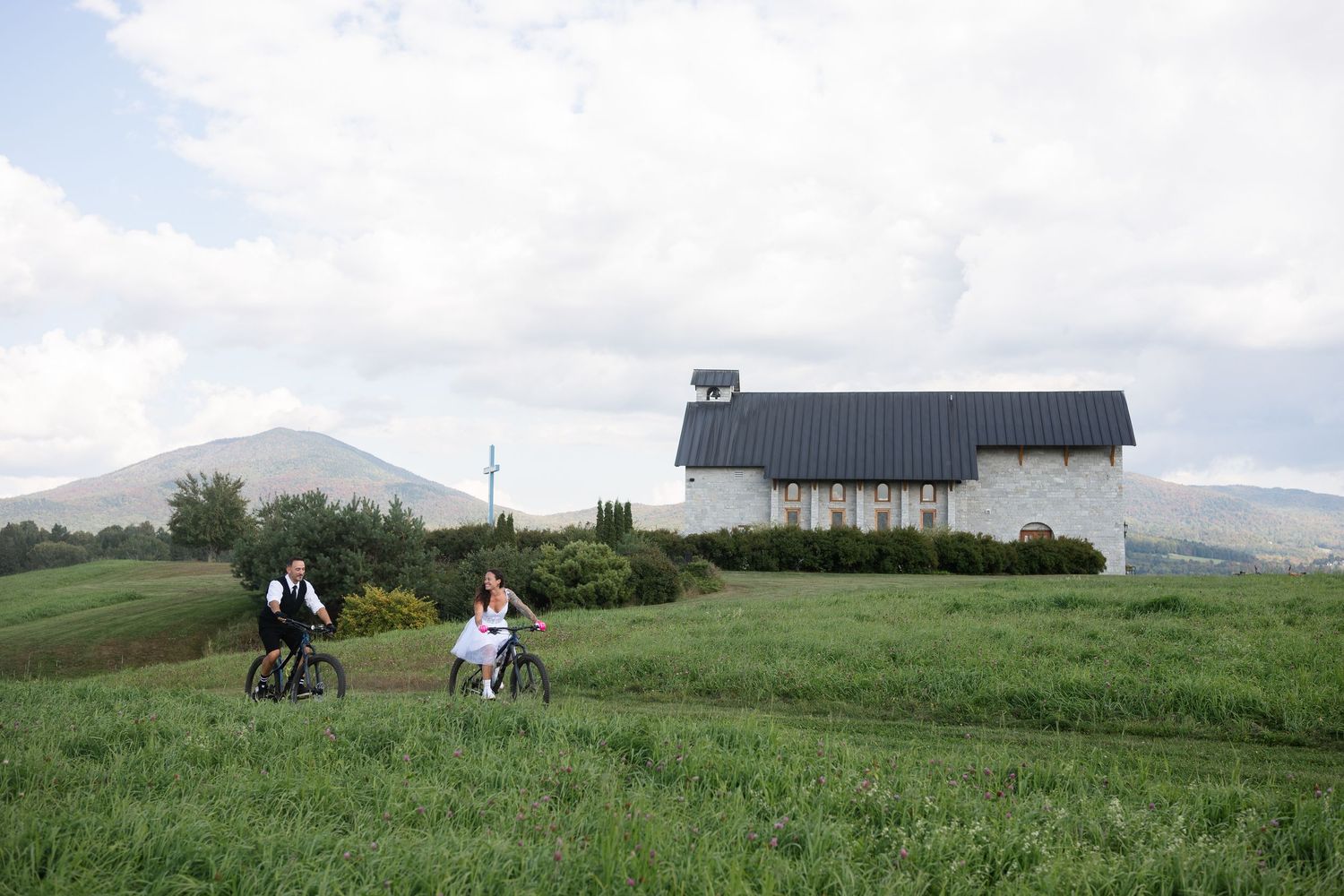 Newlywed couple rides mountain bikes through a green meadow with a rustic chapel and mountain in background.