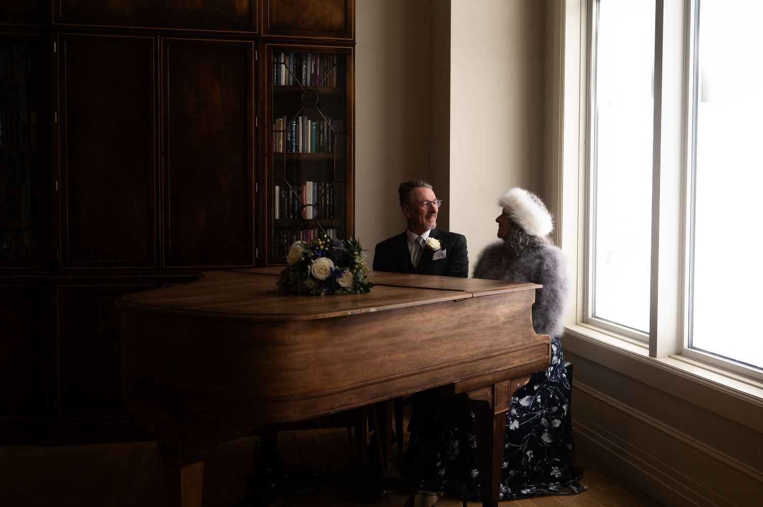 Bride and groom sharing an intimate moment at a wooden desk beside a bright window.