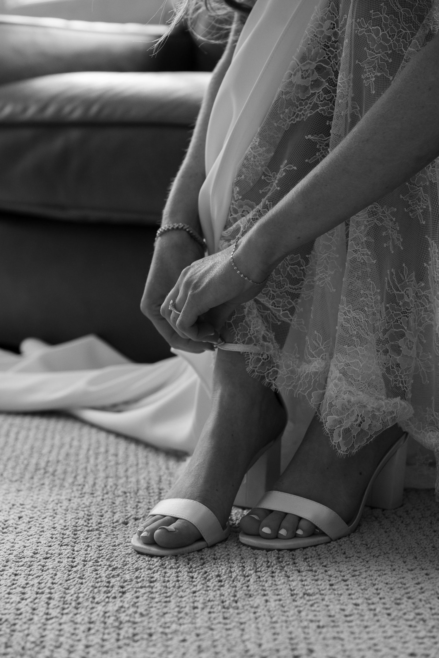 Bride adjusting her white strappy heels beneath a lace wedding dress, black and white photo.
