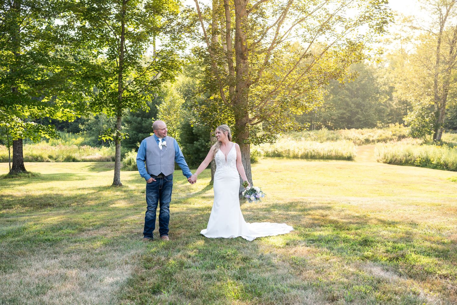 Bride and groom hold hands walking through a sunny green meadow with trees in golden light.