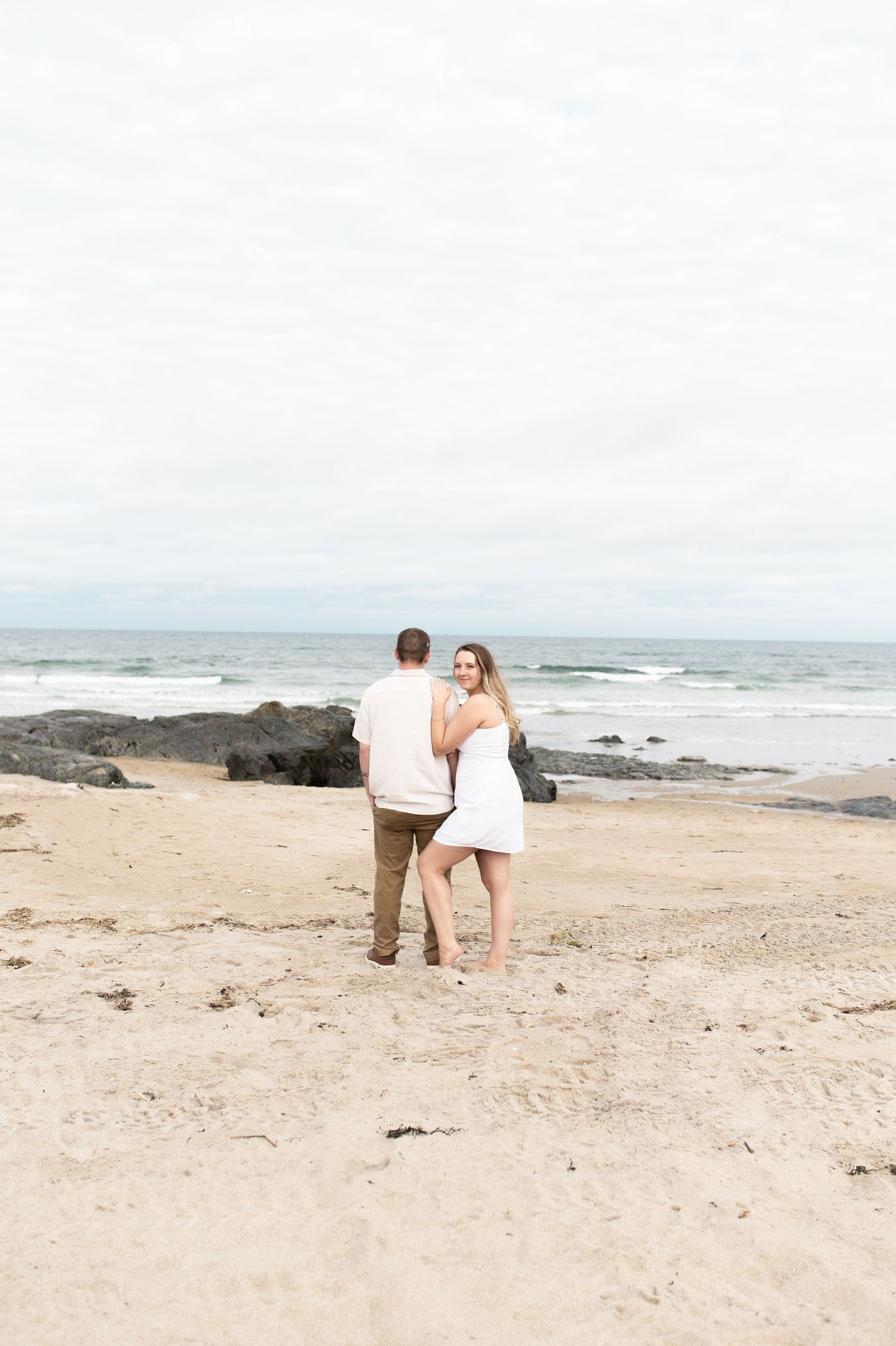 A couple walks hand in hand on a sandy beach with rocky shores and ocean waves.