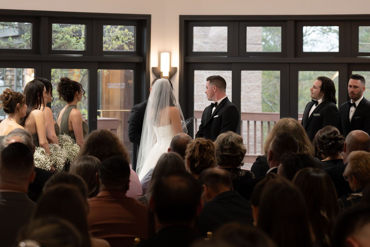 Bride and groom exchange vows at indoor wedding ceremony with guests seated in wooden chairs.